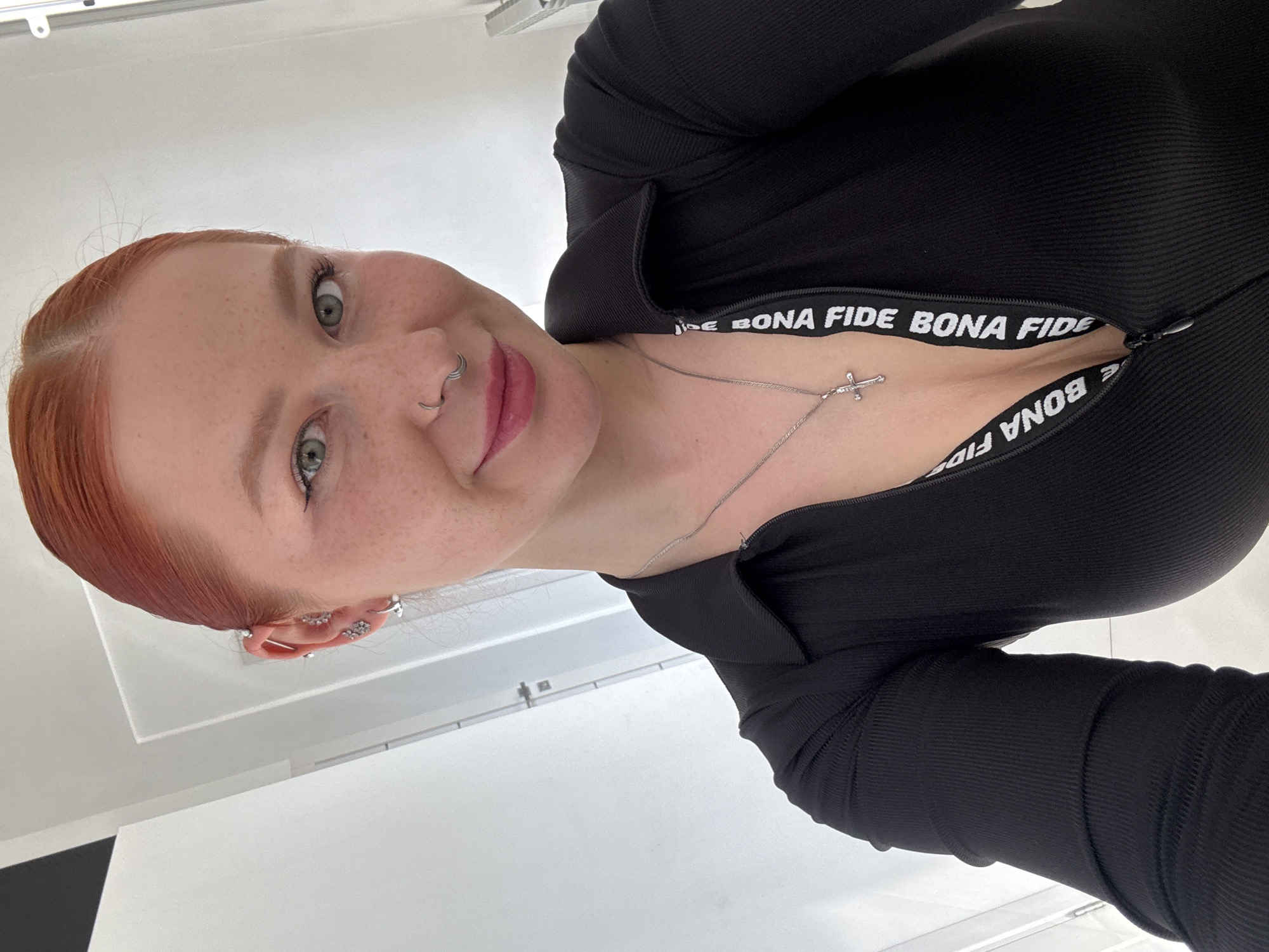 A head-and-upper-torso portrait captures a young adult female indoors, against a plain white background, consistent with a minimalist room or bathroom, in Rostov-on-Don, Russia. The subject has reddish-orange hair pulled back, fair skin with freckles, and light green eyes, looking directly at the viewer with a subtle smile. Her left ear features multiple piercings, including an industrial bar and several decorative studs, while her septum displays a stacked hoop piercing. She wears red lipstick. Her attire consists of a black, ribbed, long-sleeved collared top with a partially unzipped front. The unzipped section reveals an underlying white strap marked repeatedly with "BONA FIDE" in black uppercase letters. A silver-toned chain necklace with a small cross pendant rests on her chest.