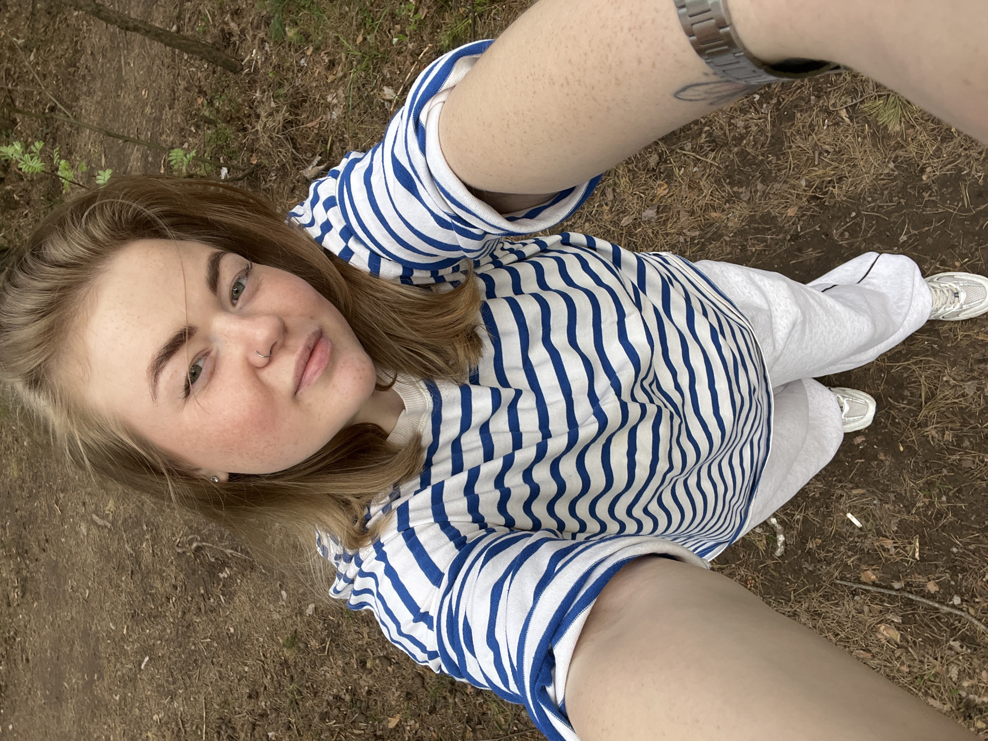 A young woman takes a selfie from an overhead perspective, seemingly outdoors. She is fair-skinned with blonde hair and wears a blue and white striped shirt and white pants. Her clothing suggests a casual setting. The ground appears to be covered in dead leaves and small twigs, indicating a wooded or park area. The presence of a wristwatch on her extended arm, as well as the lack of background buildings, suggests the photo was taken in a natural environment, possibly a park near the city of Chaikovsky, Russia. There are no apparent interactions with other individuals.