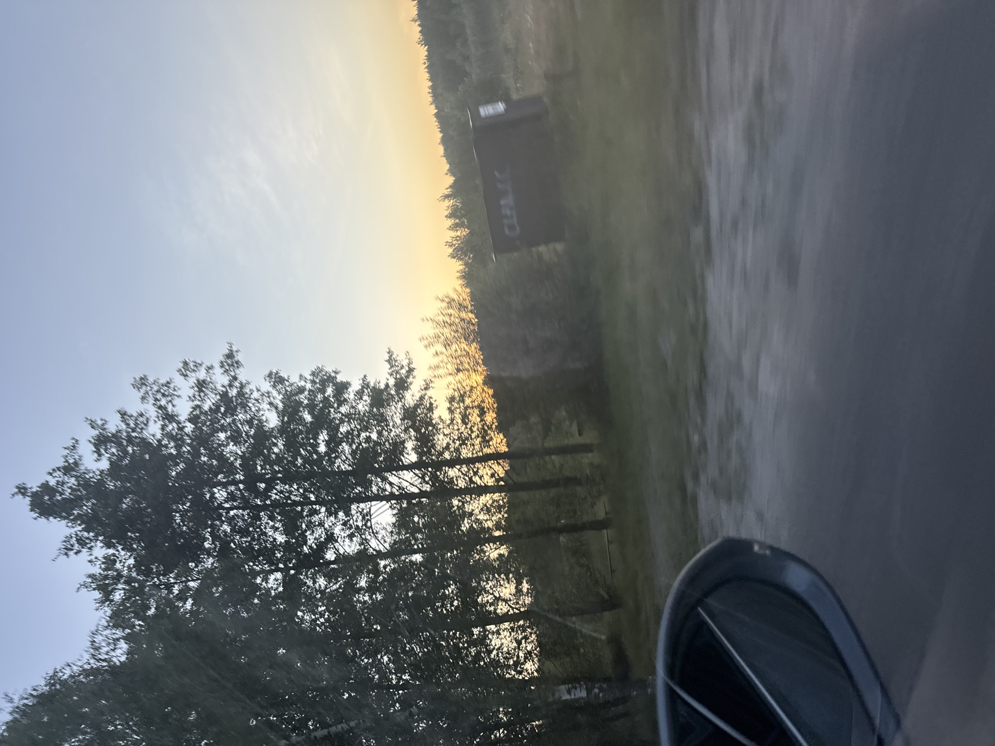 The image depicts a roadside scene in Orlivka, Ukraine, likely captured from a moving vehicle. The sky is a pale blue, transitioning to a warm, golden hue near the horizon, indicating either sunrise or sunset. Several trees with dark green foliage are visible on the left, their branches silhouetted against the bright sky. In the background, a dark, rectangular structure with white text is situated amidst a wooded area. The text on the structure appears to read "CULIAC." The ground to the right is a gray, textured surface, possibly asphalt, with a blurred appearance suggesting motion. The lower right corner of the image shows a portion of a dark, rounded object, which is likely a side-view mirror of the vehicle. The overall mood is serene, with the soft light of dawn or dusk casting a peaceful glow over the landscape. The presence of the trees and the distant structure suggest a rural or semi-rural setting.