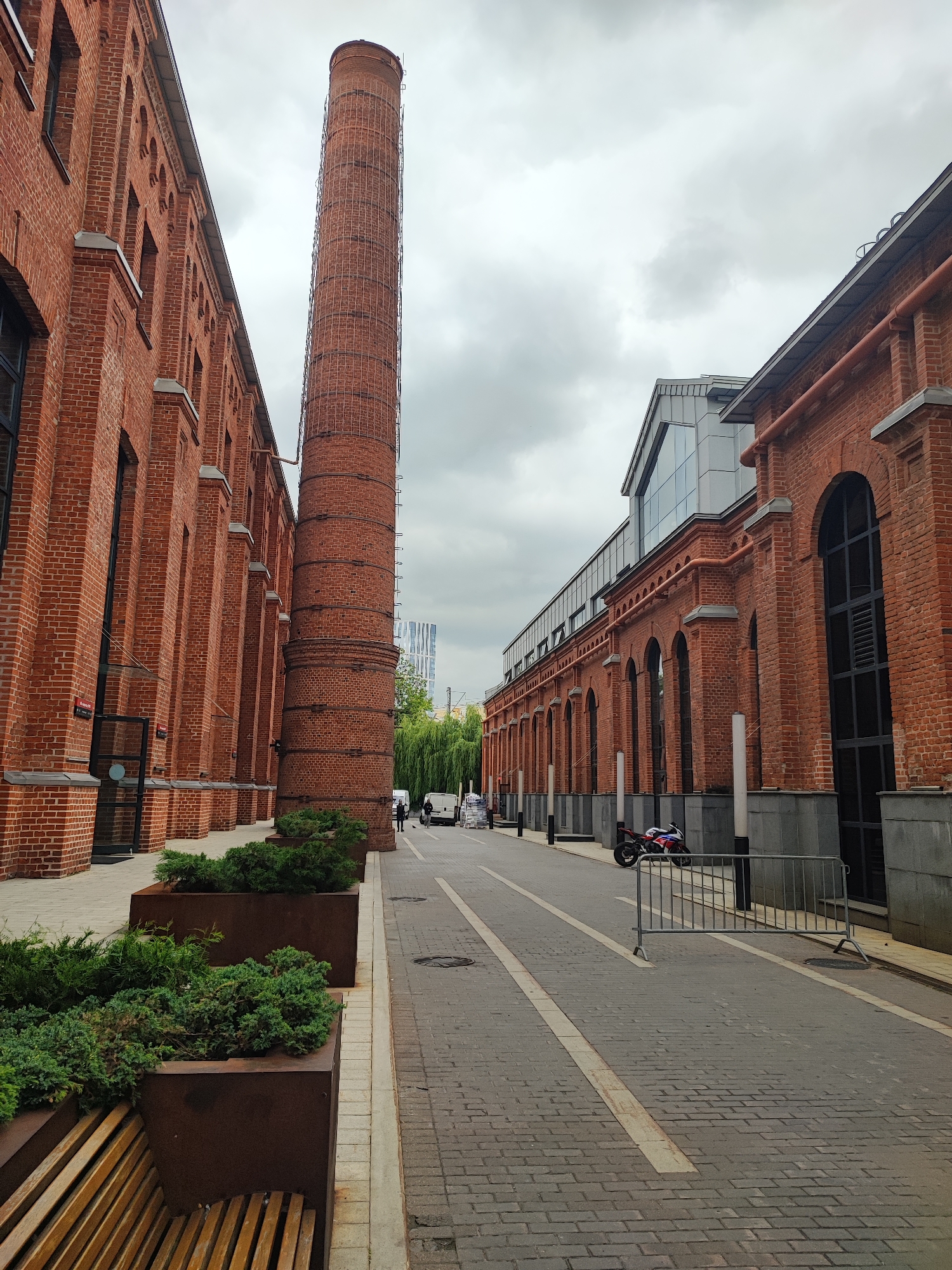 This image depicts an outdoor urban scene in Moscow, Russia, likely in a redeveloped industrial area. The setting features two long, red brick buildings with tall, arched windows and a distinctive industrial-era chimney stack. The buildings have a modern renovation, indicated by a section of glass and metal architecture integrated into the roofline of the building on the right.
In the foreground, a pathway paved with grey bricks is lined with large, dark metal planters containing green shrubs and a wooden bench. The path is bisected by white lines. A metal barrier is visible further down the path.
On the right side of the image, parked near the building, is a red, white, and blue motorcycle. Further down the path, two people are visible, along with two white vans, suggesting some ongoing activity or a place with foot and vehicle traffic. The overall atmosphere is somewhat subdued, likely due to the overcast sky. The time of day is indeterminate, but the cloudy weather suggests it's not bright sunlight. The scene conveys a sense of historical architecture being repurposed for modern use, creating a unique urban environment. There is no visible text in the image.