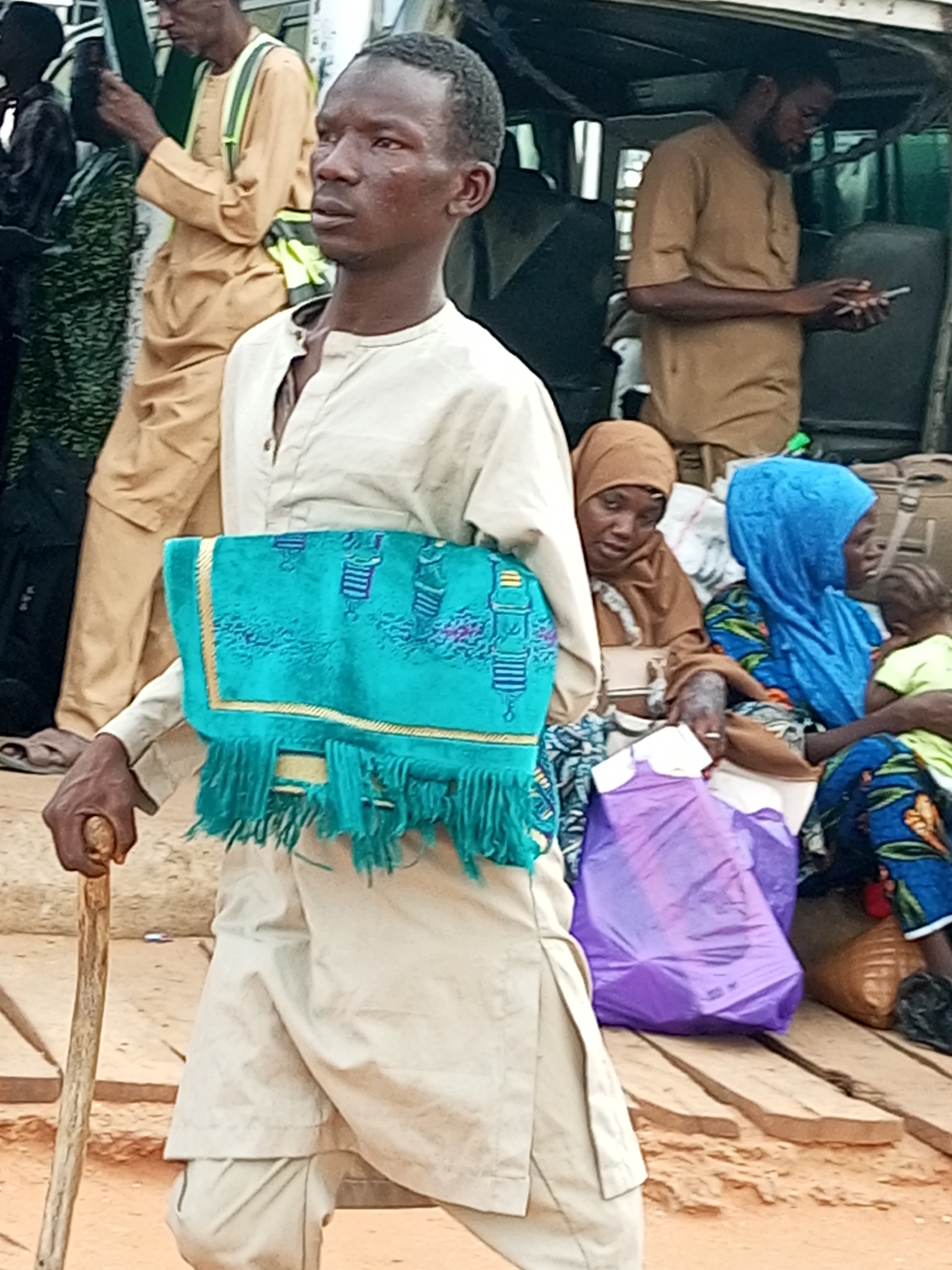 In Gombe, Nigeria, a man stands in the foreground holding a folded turquoise cloth with gold trim and fringes. He is wearing a light-colored tunic and trousers and is leaning on a wooden walking stick. Behind him, other people are visible, some seated on wooden planks and others standing. One woman wears a brown hijab, and another wears a blue hijab. A small child is being held by the woman in the blue hijab. Luggage, including a purple plastic bag and brown and black bags, is scattered around. The environment appears to be an outdoor area with a sandy ground and wooden structures.