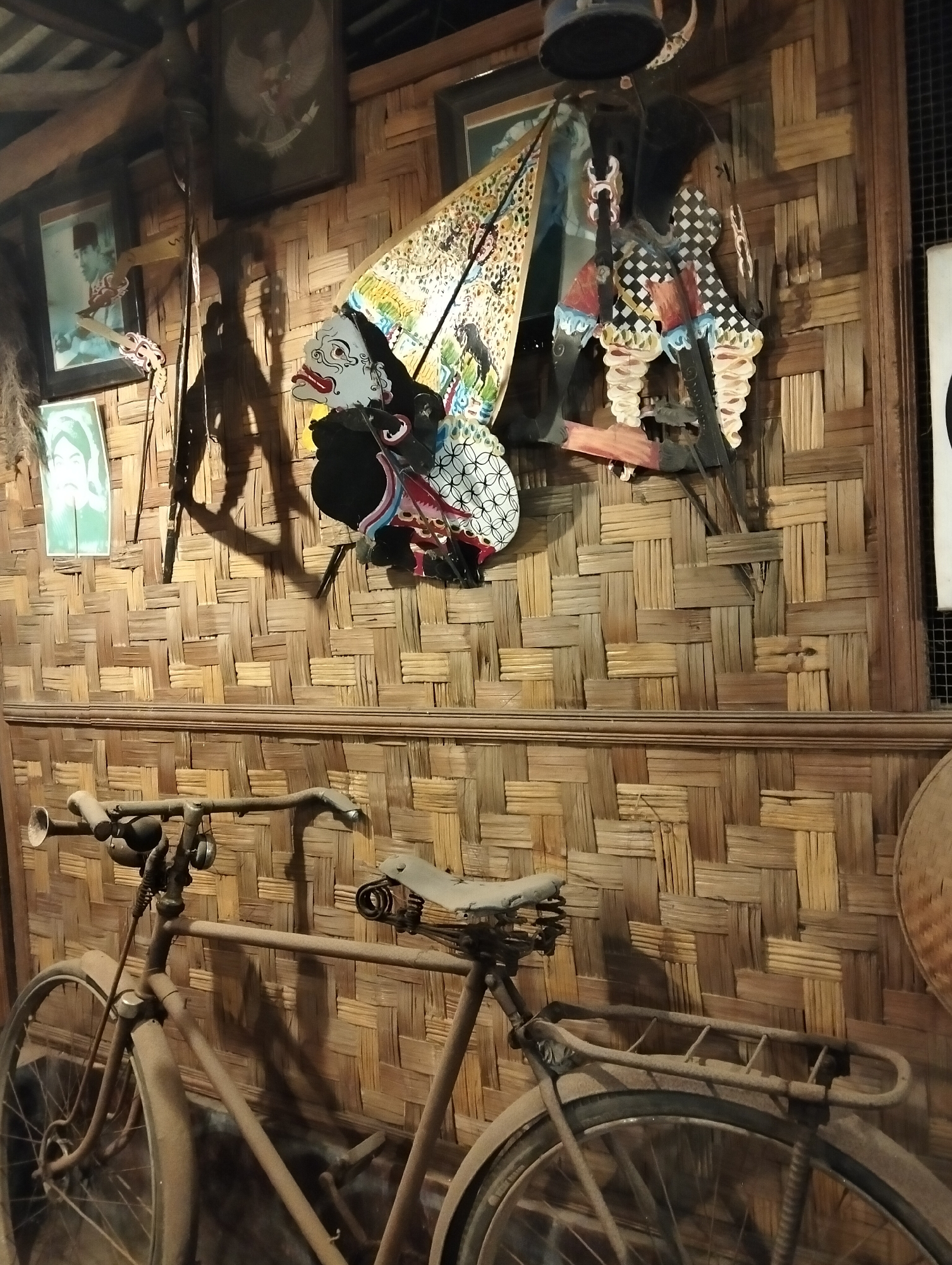 In Kedungrejo, Indonesia, a room features a wall covered in woven bamboo. On this wall, a framed image of Garuda Pancasila is displayed at the top center, to the left of it are two framed portraits, one in black and white and another colored. Below these, a traditional Javanese Wayang Golek puppet is displayed, depicting characters with detailed facial features and costumes. To the right of the Wayang Golek puppet, another framed picture is visible. In the foreground, a vintage, rusty bicycle is parked. The bicycle is equipped with a horn, two bells, and a sprung saddle, with a luggage rack over the rear wheel. The overall scene evokes a sense of cultural heritage and historical preservation.