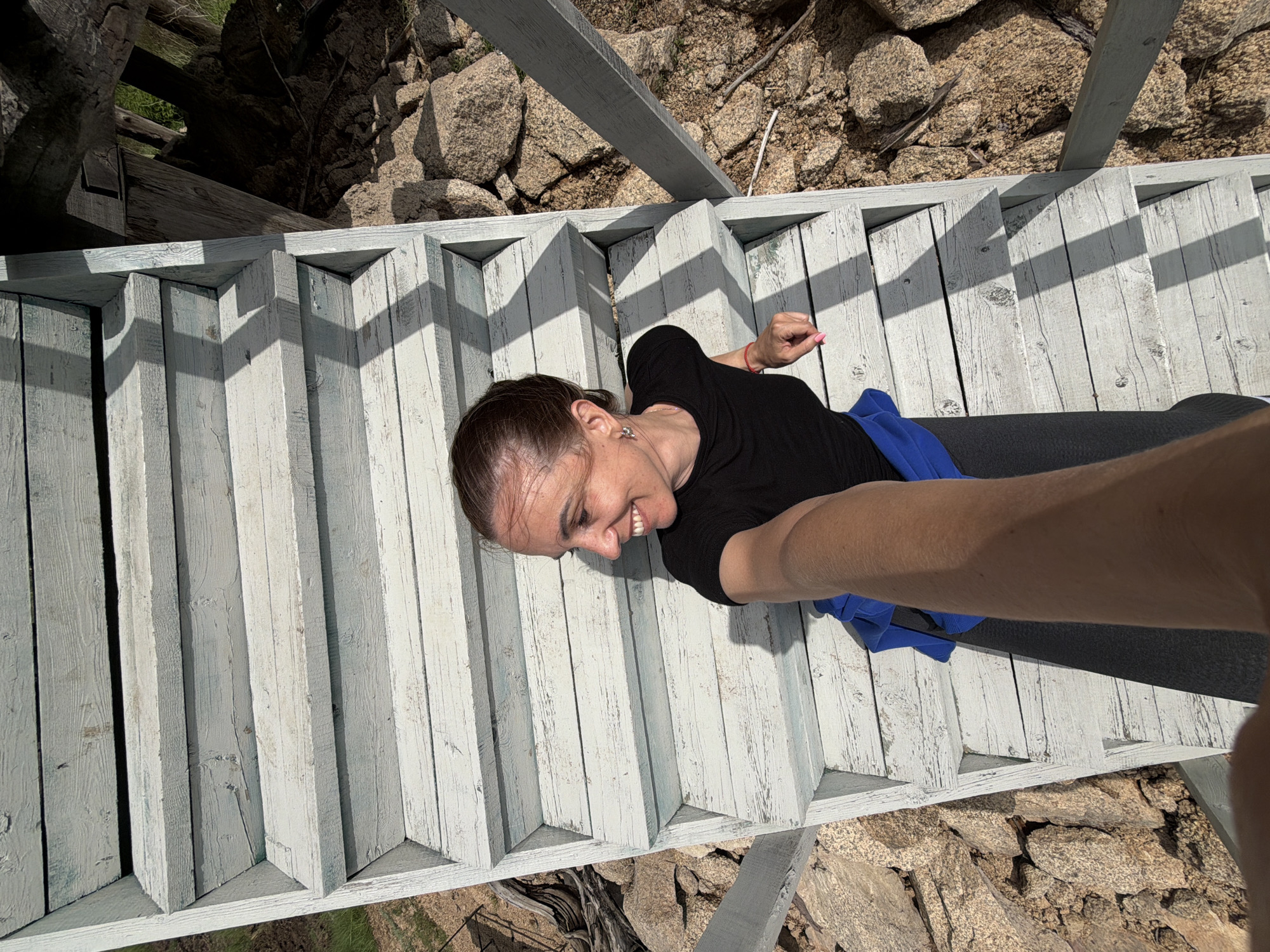 In an outdoor scene, a woman with brown hair pulled back smiles and looks to her right as she takes a selfie on a weathered, light blue wooden staircase. She is wearing a black t-shirt and blue shorts. The staircase is surrounded by large, light brown rocks and sparse vegetation. Sunlight casts shadows on the stairs, creating a striped pattern. The woman's arm and hand, holding the camera, extend into the foreground of the image. The location is identified as Unknown, Russia.