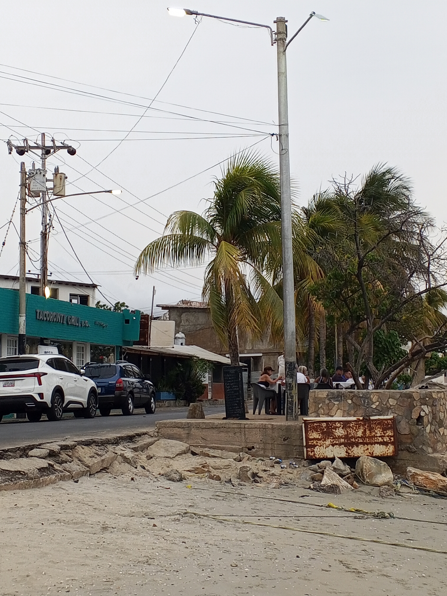 The image shows a street scene in Juan Griego, Venezuela. A turquoise building with "Tacoronti Grill" on its facade is visible on the left, with two parked vehicles (a white SUV and a dark blue SUV) in front of it. Overhead power lines and a utility pole are visible.
On the right side of the street, there is a stone retaining wall with people seated at tables. The people appear to be dining. Palm trees and other vegetation are also present. In the foreground, there is a sandy area with debris, a rusted metal box, and broken concrete.