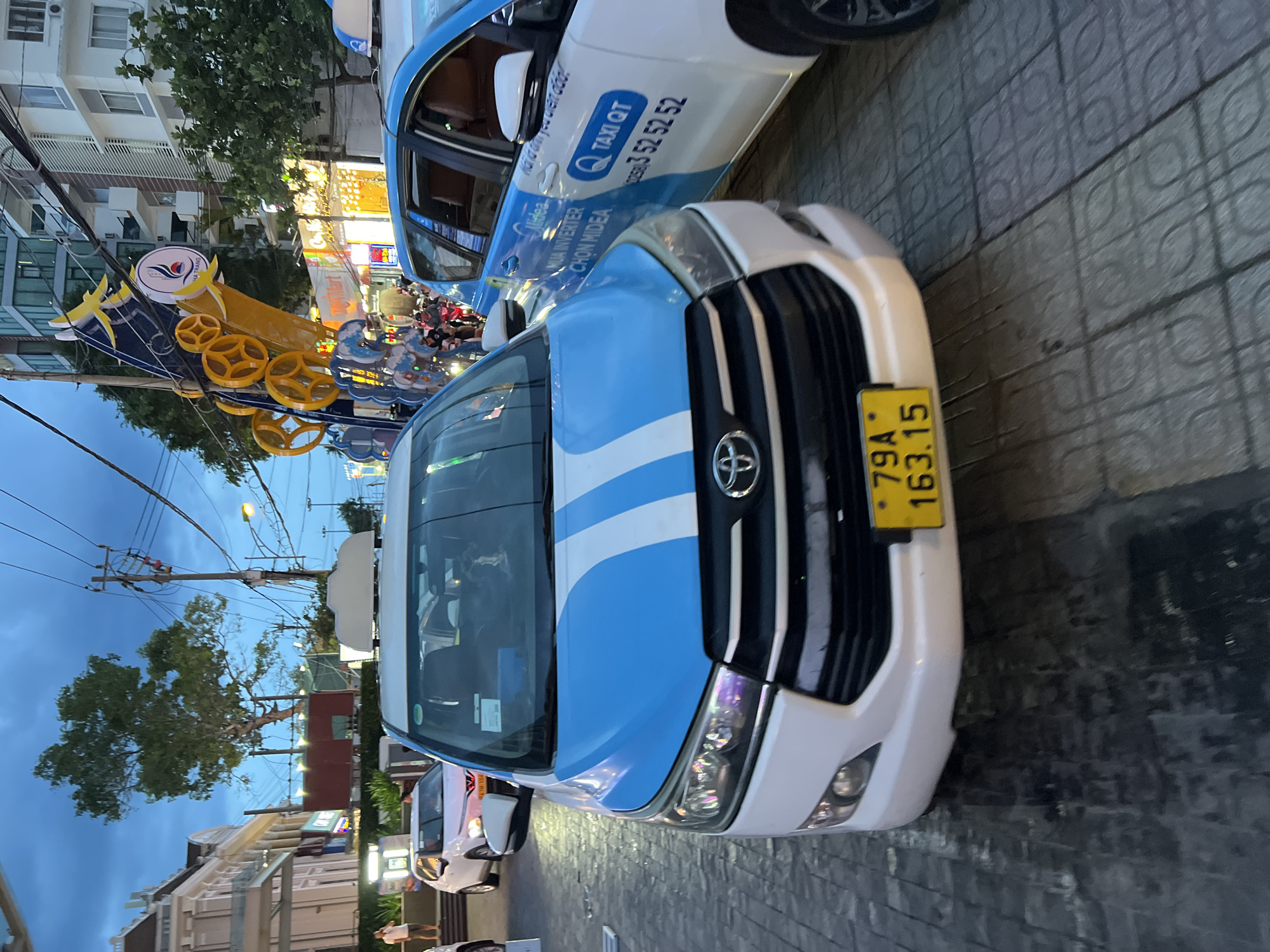 The image depicts a street scene in Nha Trang, Vietnam, at dusk. In the foreground, a white and blue taxi, identified as a Toyota Innova, is parked. The taxi has a yellow license plate with the characters "79A 163.15". To the right of this taxi is another white taxi with blue branding, displaying "TAXI QT" and a phone number. The street is paved with textured tiles, and a wet sheen suggests recent rain.
In the background, a large, brightly colored decorative structure, possibly a temporary installation for an event, is visible. It features yellow geometric shapes and a blue and yellow bird-like motif with the text "NHA TRANG" on a white circular emblem. Adjacent to this structure are modern buildings with multiple stories, illuminated windows, and balconies. Trees with full foliage are scattered throughout the scene. The sky is a deep blue, characteristic of twilight, with visible power lines and streetlights.
Other vehicles, including a white car, and motorcycles, are present in the midground, indicating moderate traffic. A few individuals are visible, including a person standing near the white car on the left and others on motorcycles. The overall impression is of a busy urban environment transitioning into the evening.