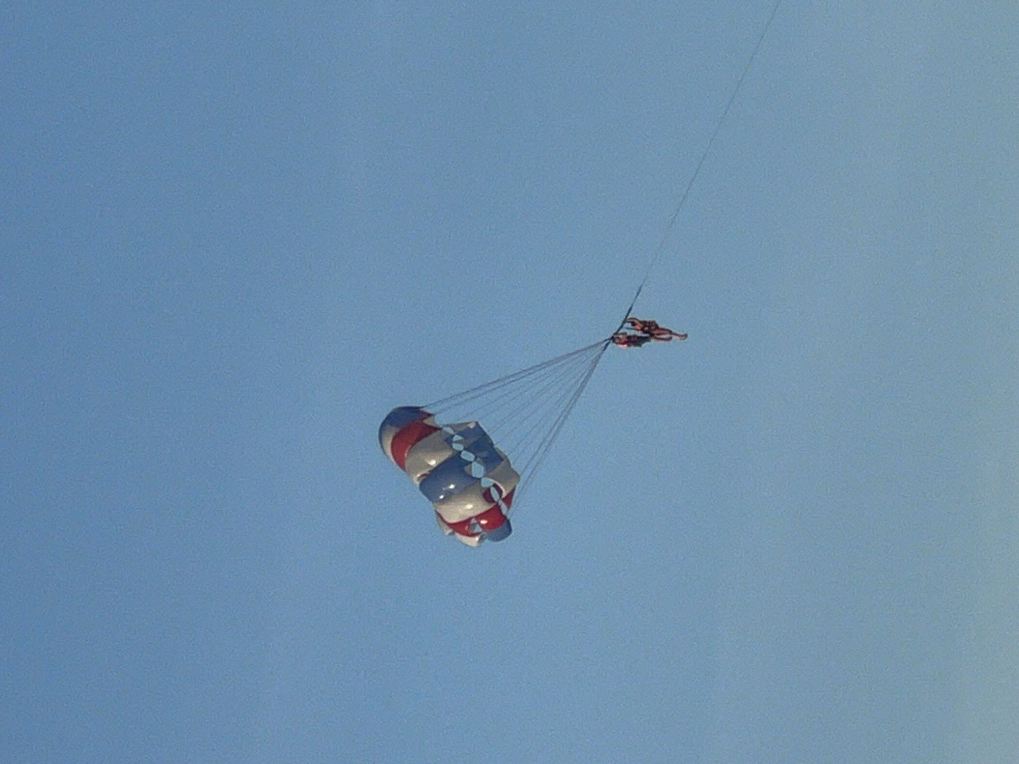 This image captures a bright, clear blue sky over the city of Đà Nẵng, Vietnam, where two individuals are parasailing. The parasail, a large canopy with alternating red, white, and blue sections, is inflated and suspended high in the air. Attached to the parasail by multiple lines is a harness system from which two people are hanging. Both individuals are dressed in swimwear and appear to be enjoying the activity. The scene suggests a recreational pursuit taking place over a coastal area or a body of water, common in Đà Nẵng.