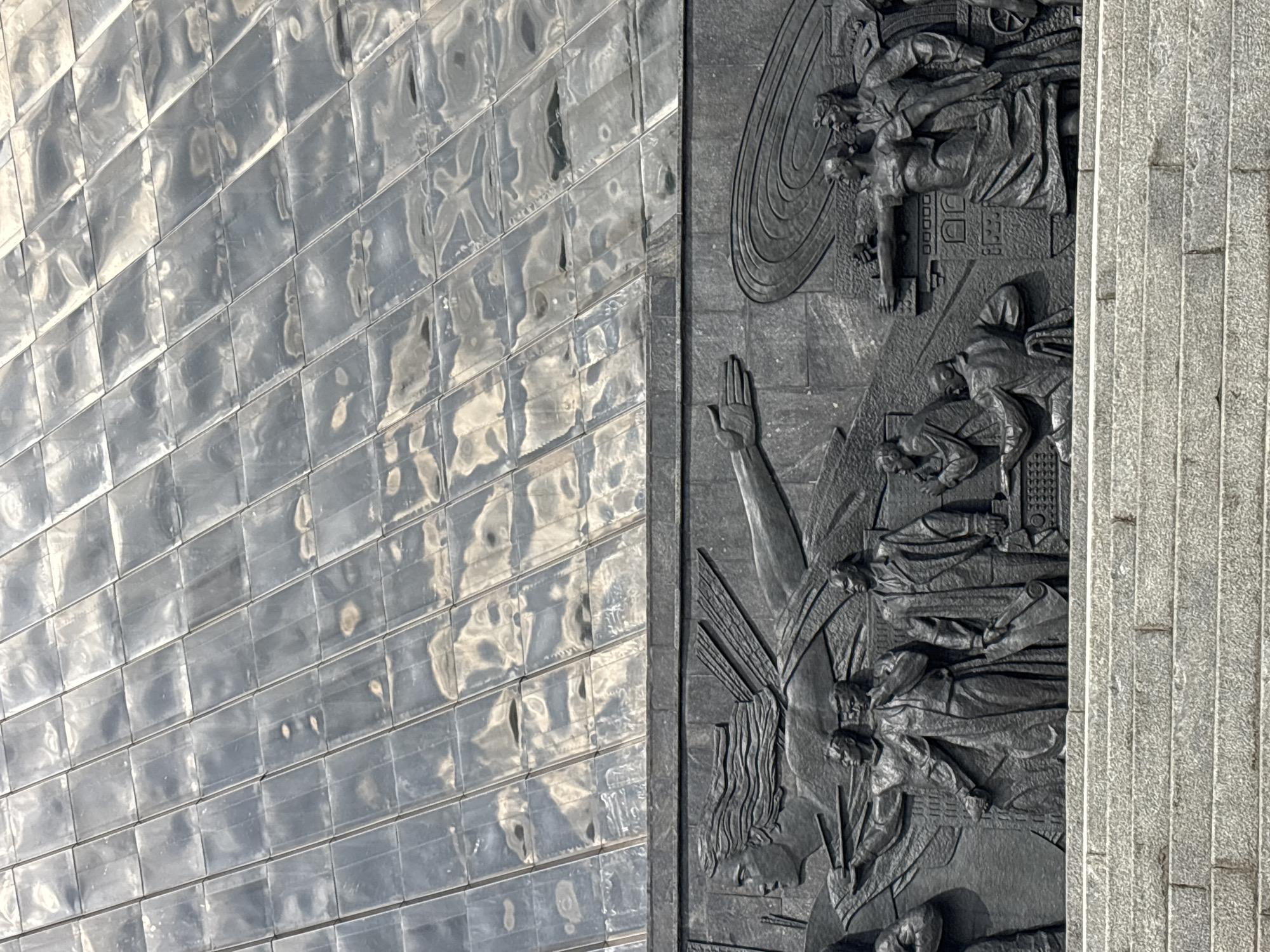 The image shows a portion of the Monument to the Conquerors of Space in Moscow, Russia. The left side depicts a titanium-clad upward-sloping structure composed of rectangular tiles. To the right is a dark-colored relief depicting figures, including a person with a raised hand and other figures in various poses. The relief extends to a section of vertically-aligned light gray stone.
