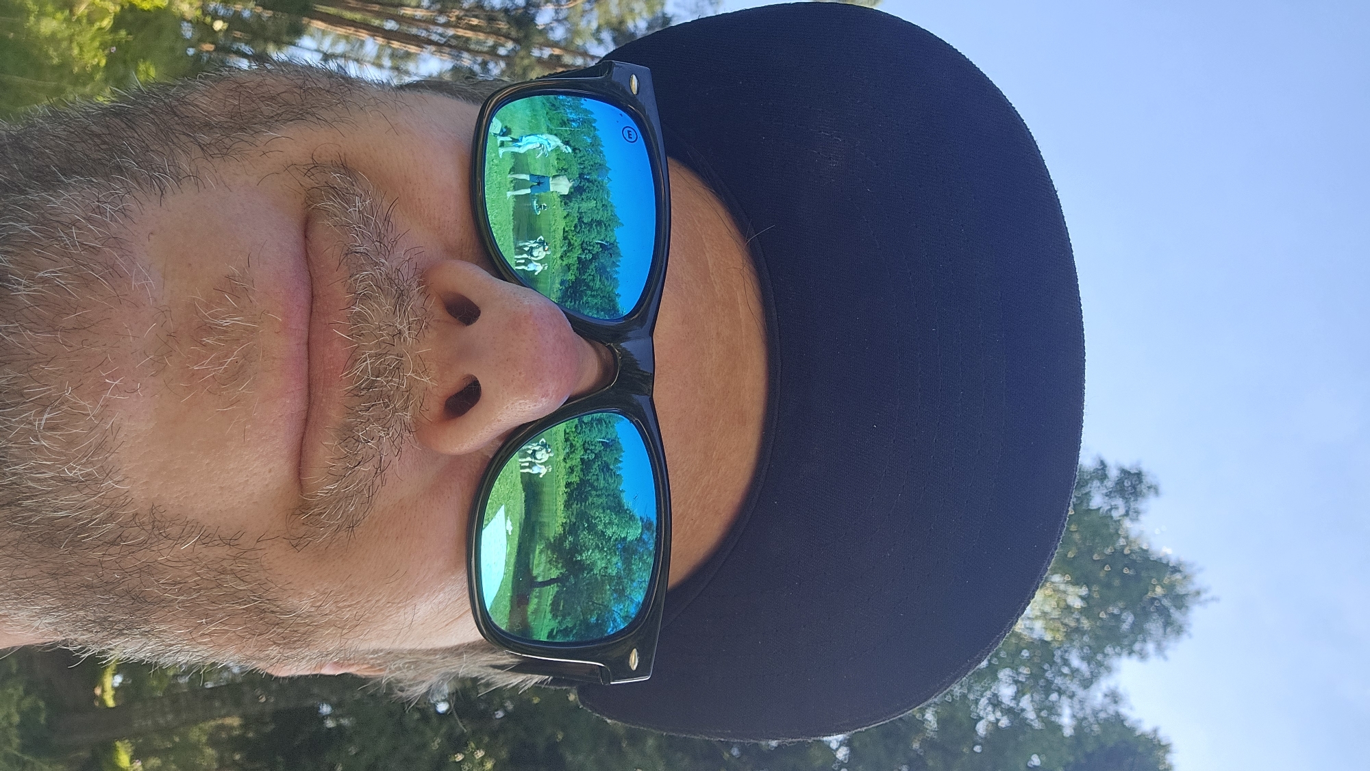 A close-up, low-angle selfie shows a person from the nose down, wearing a black baseball cap and black-rimmed sunglasses. The sunglasses' mirrored lenses reflect a scene of people gathered near a body of water, surrounded by trees and grass under a clear blue sky. The environment appears to be a natural outdoor setting, possibly a park or a lakeside area. The person in the foreground has a light-colored beard and mustache. The image is taken outdoors, with trees visible in the background on either side of the frame. The location within Voinskaya Chast' Mchs, Russia, is not discernible from the image itself.