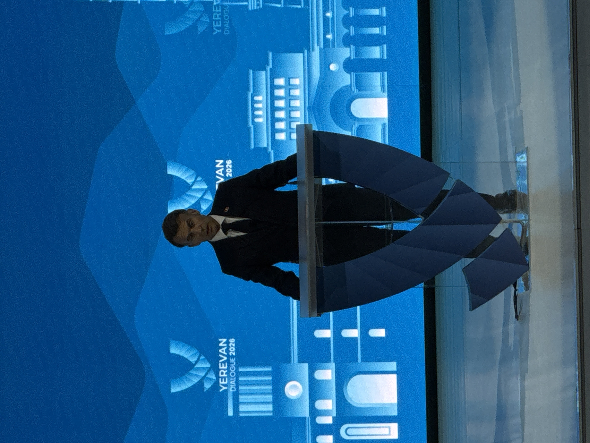A man in a dark suit and tie stands at a podium with a blue, geometric design. The background features a graphic with the words "YEREVAN DIALOGUE 2026". The location is unknown.