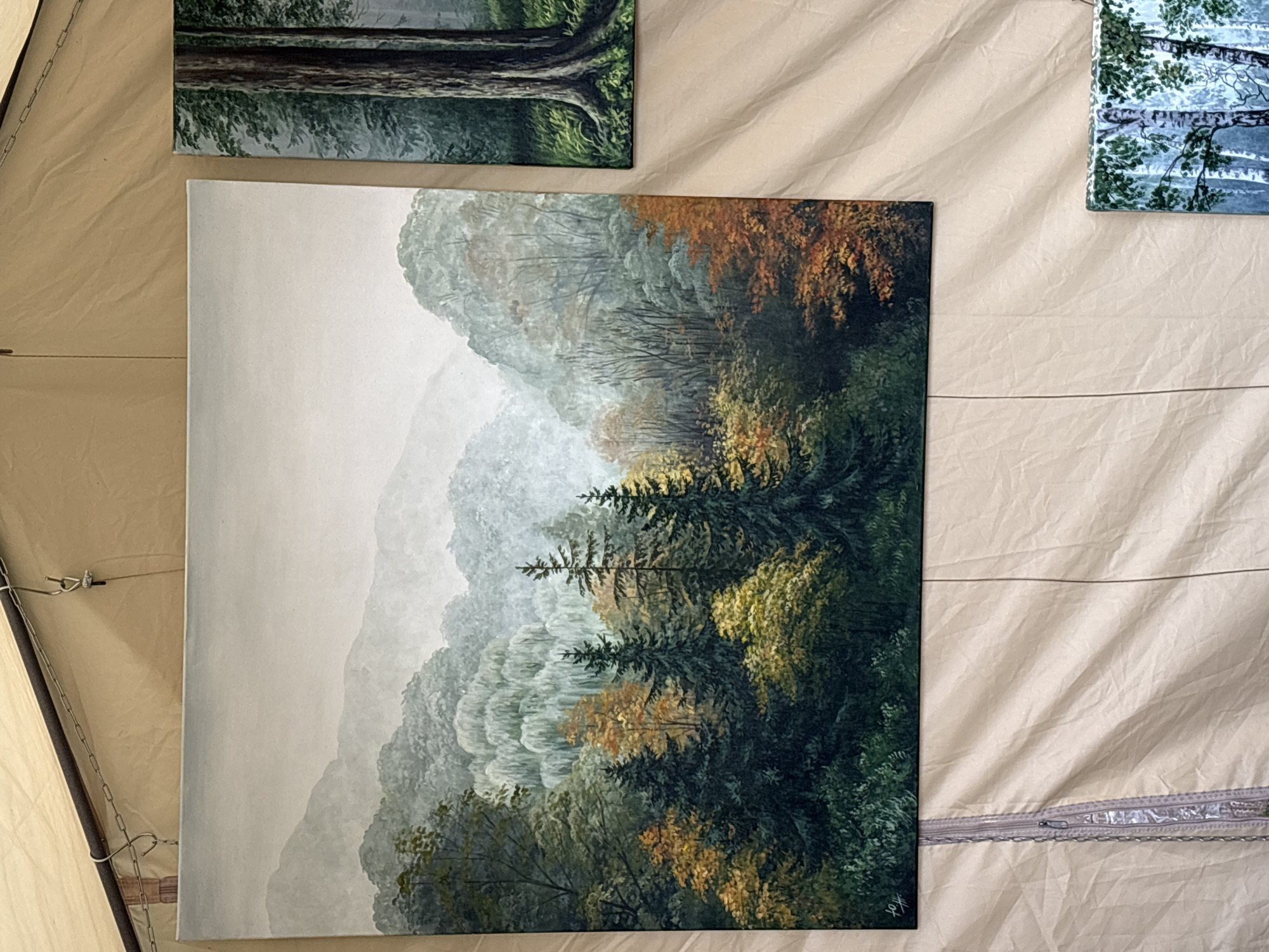 The image shows three paintings hanging on a light-colored fabric backdrop, possibly inside a tent or market stall. The main painting in the center is a landscape of a misty forest with rolling hills in the background. The trees in the foreground display a mix of dark green and autumnal colors, with some lighter yellow and orange foliage. The painting on the right is a close-up of tree trunks in a forest, with dappled light filtering through the leaves. The smallest painting at the bottom right appears to be a birch forest scene. The paintings are hung using metal hooks and chains from a dark rod at the top. The backdrop is creased and textured, suggesting it might be canvas or a similar material. There are no people or animals visible in the image. The location is not specified within the image itself.