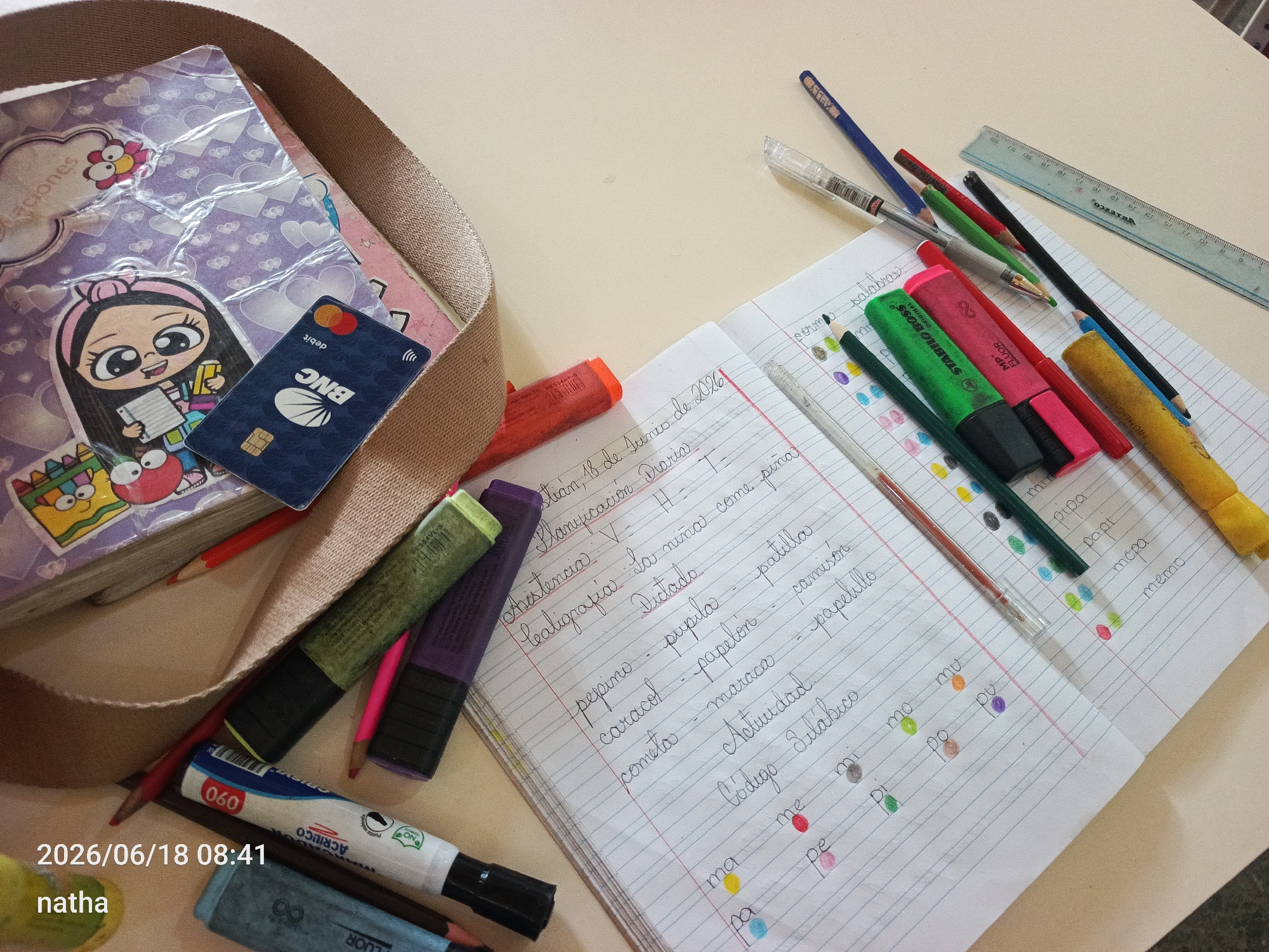 A still life image captures a collection of school supplies and personal items arranged on a light-colored surface, possibly a desk. The scene is lit by natural light, suggesting daytime. The items include a notebook with handwritten notes and drawings, several colored pens and pencils, a ruler, a debit card, and what appears to be a toiletry bag or backpack. The notebook, open to a page with red and blue lines, contains Spanish text and colorful dots. Some of the visible words include "Dios", "Planificación", "Asistencia", "Caligrafia", "Dictado", "Cometa", "Pepino", "Caracol", "Naturaleza", and "Sílabas". A debit card from BNC is also visible. The overall impression is one of preparation for school or study.
A timestamp in the lower left corner indicates "2026/06/18 08:41" and the name "natha" is present below it. The presence of "San Sebastián, Venezuela" as location context suggests this is where the image was taken. No weather information is available.