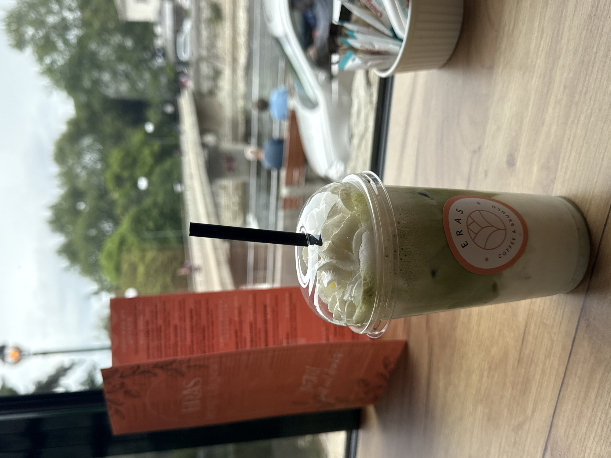 A close-up photograph shows a table inside a cafe. On the table is a clear plastic cup filled with a green drink topped with whipped cream and a black straw. The cup has a circular logo on its side with the words "ERAS" and "COFFEE & BRUNCH" around a stylized leaf design. Next to the cup is a reddish-orange menu card. In the background, visible through a window, is an urban scene with trees, buildings, and vehicles, partially obscured by reflections. This photo was taken in Asenovgrad, Bulgaria.