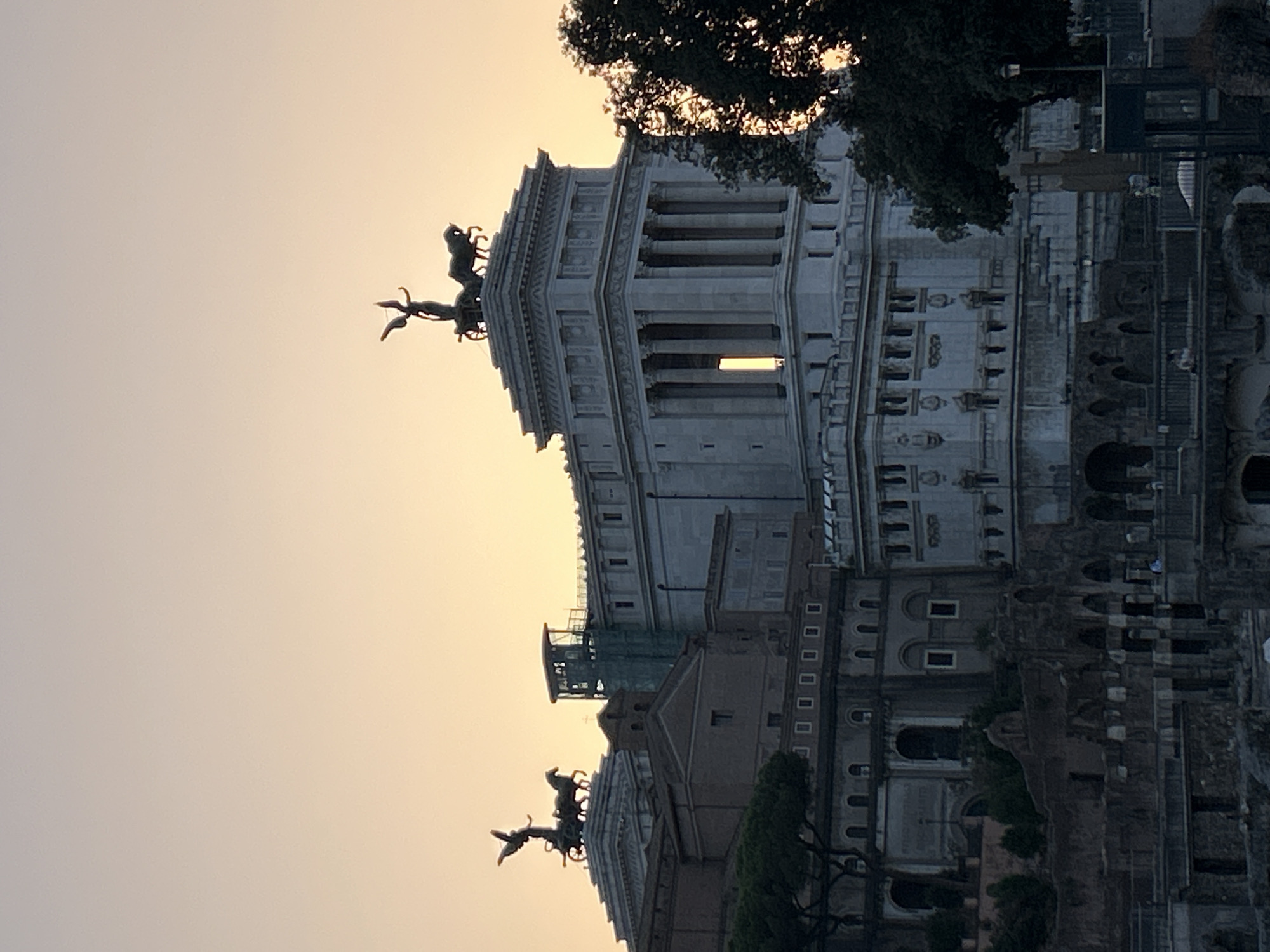 The image captures a view of the Altare della Patria, also known as the Victor Emmanuel II Monument, in Rome, Italy. The monument, a grand neoclassical structure made of white marble, dominates the frame. It features a large central edifice with columns, topped by a quadriga (a chariot drawn by four horses) with a statue of the goddess Victoria. To the left, another similar monument with a quadriga is visible on a smaller building. The sky above is a soft gradient of orange and yellow, suggesting either sunrise or sunset. In the foreground, ancient ruins, likely part of the Roman Forum, are visible, with stone structures and arches. Trees, one of which is silhouetted against the bright sky on the right, add to the scene. The lighting suggests a late afternoon or early evening time.