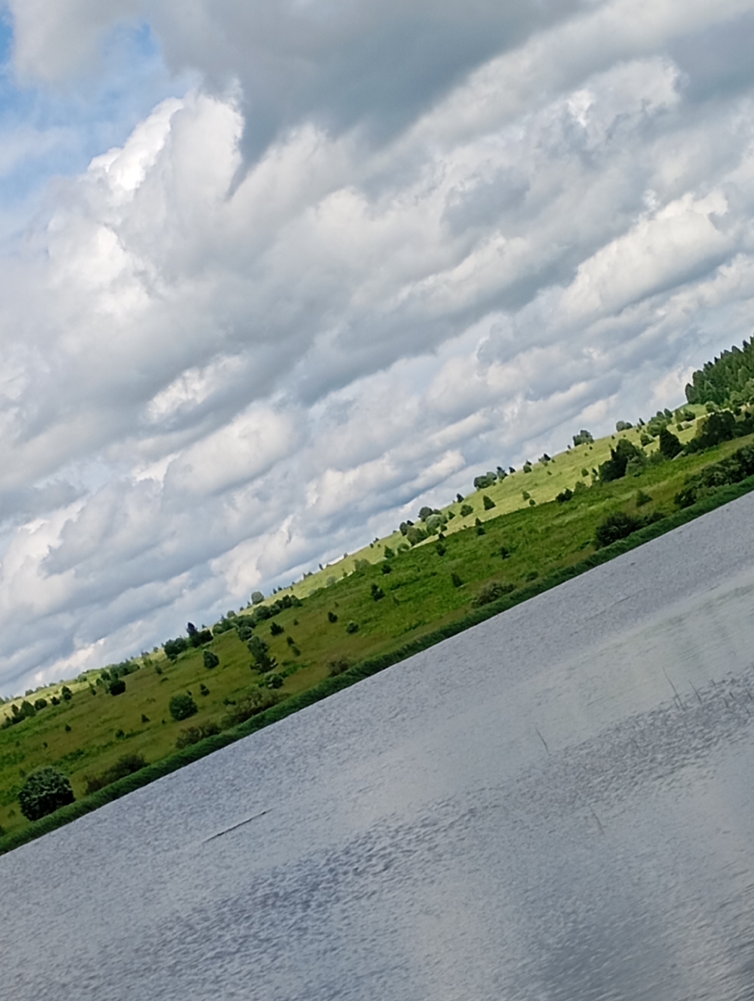 The image shows a body of water in the foreground, with a green, grassy hillside dotted with trees in the middle ground. The hillside is under a sky filled with dramatic, grey and white clouds. The scene is tilted, suggesting it was captured at an angle. The presence of a body of water, a grassy hillside with trees, and a cloudy sky indicates a natural, outdoor environment. Given the context of Рябки, Russia, this scene likely depicts a rural or semi-rural landscape near a body of water. There are no people or specific objects of human construction clearly visible beyond what appears to be a line of trees at the top of the hill and possibly some thin vertical structures in the water.