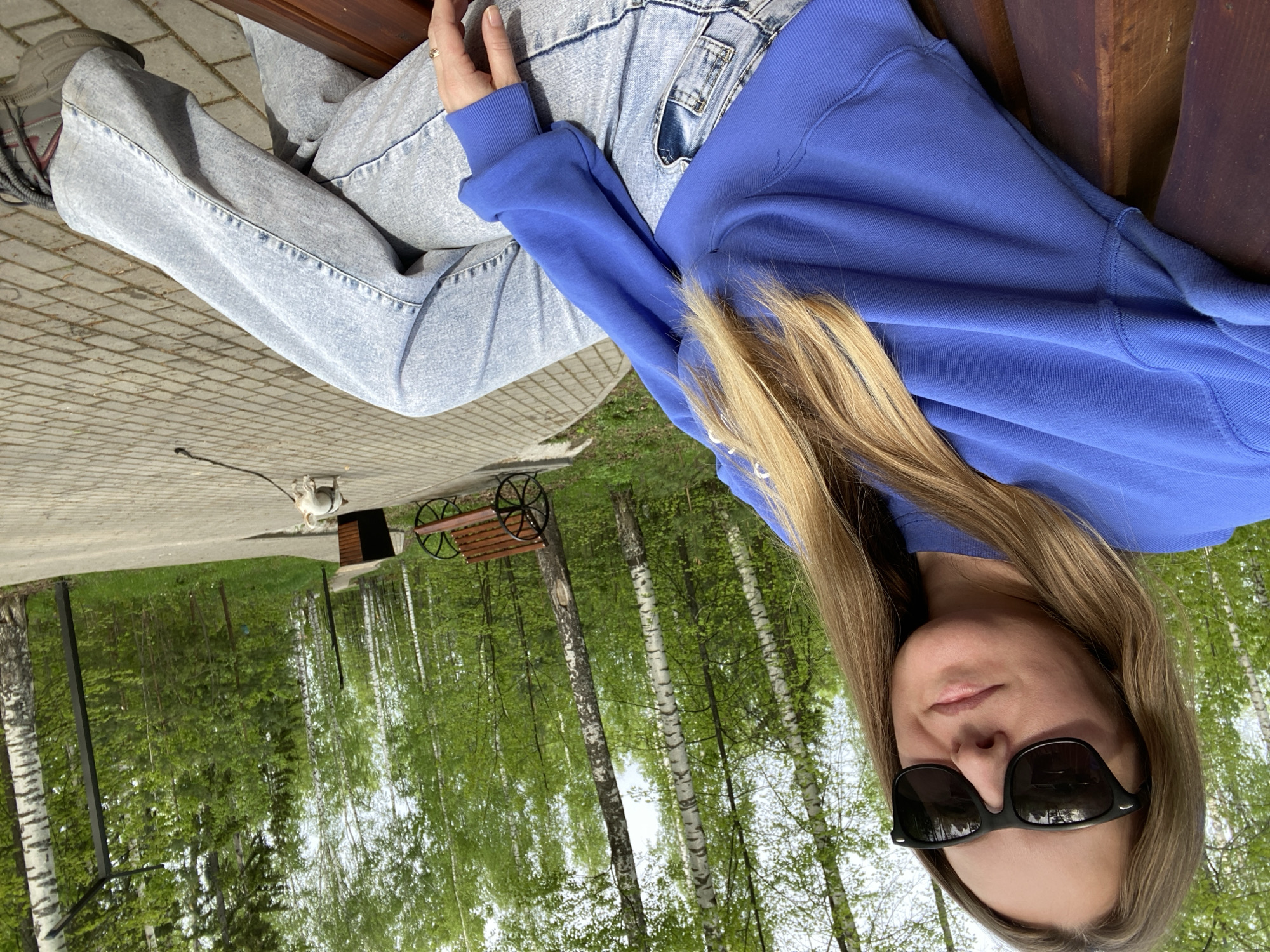 A woman with blonde hair and sunglasses is sitting on a bench, wearing a blue sweatshirt and light blue jeans. The scene is outdoors, likely in a park or recreational area, evidenced by the trees and the brick-paved walkway. A small dog is visible in the background, leashed. The location is Noviy, Russia, as indicated by the prompt. The woman appears to be resting or relaxing on the bench.