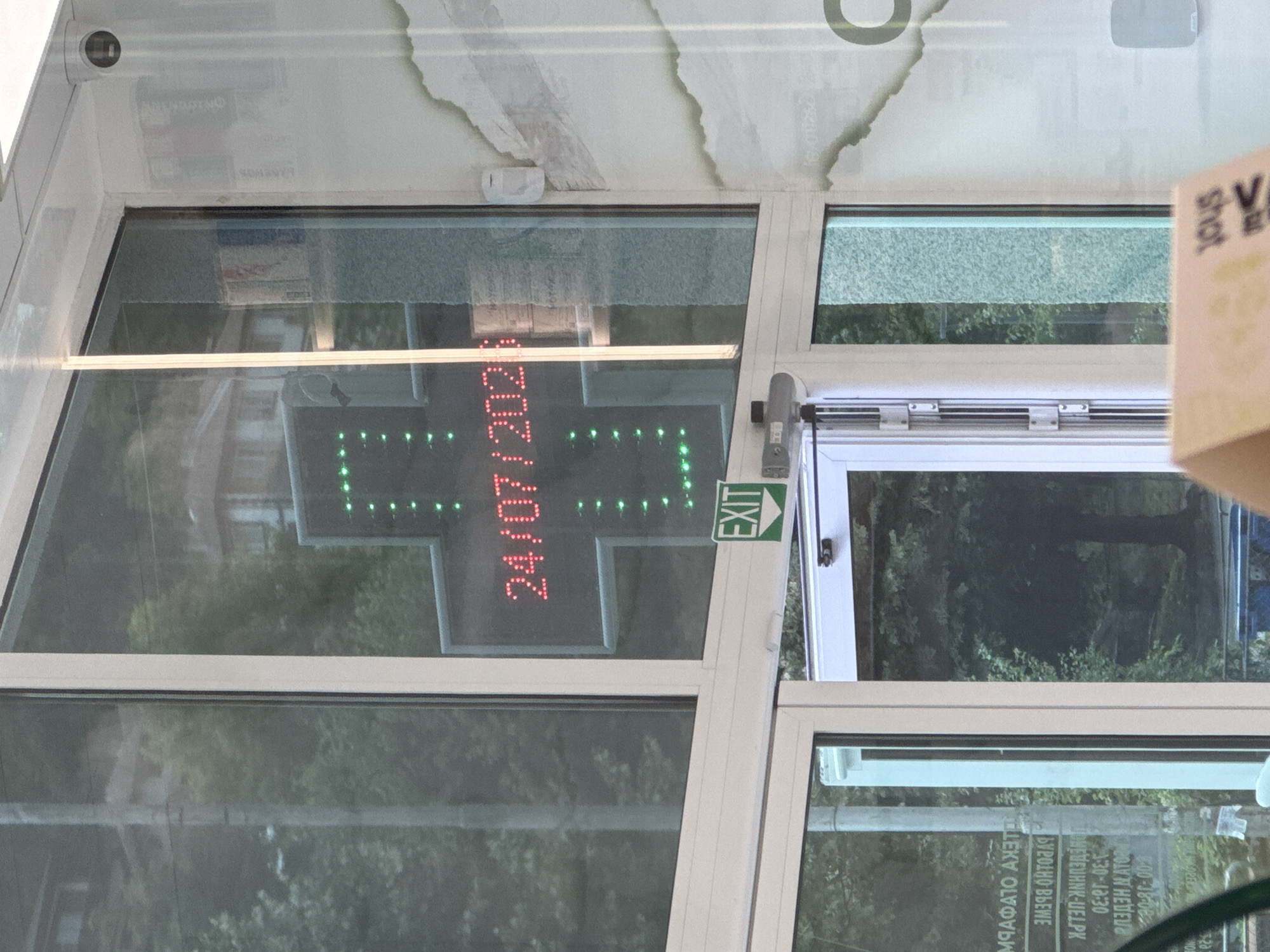 The image shows the exterior of a pharmacy with a digital cross-shaped sign displaying the date "24/07/2023" in red LED lights. A green "EXIT" sign is visible below the digital display. The pharmacy appears to be located on a street in Madan, Bulgaria, as indicated by Cyrillic text on a reflection in the window, which reads "МАФАТО АХЕТО" and "3M938 OHTO8A9", likely representing pharmacy names or information. The building has large glass windows and doors, with reflections of trees and buildings visible. There are no people clearly visible in the image.