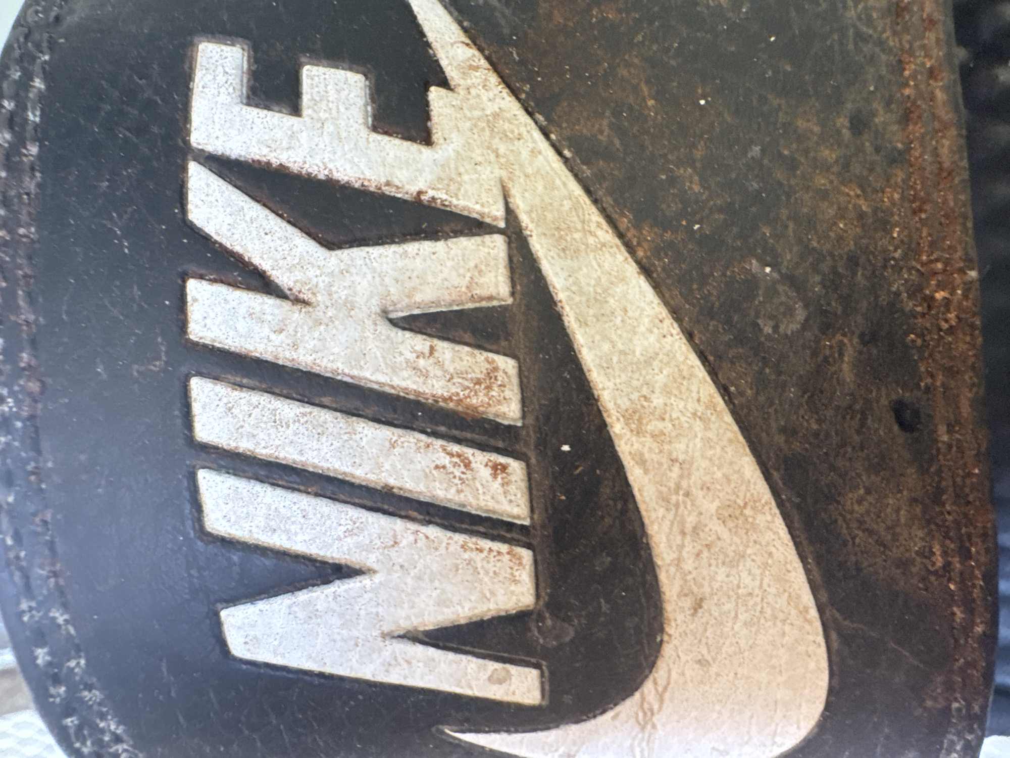 The image shows a close-up of a black Nike logo with a white swoosh on a dark, textured surface, possibly a shoe or slide. The logo appears weathered and dirty, with visible scuffs, scratches, and reddish-brown discoloration. The background is dark and out of focus. The image does not provide any information about the city of Zgrade, Montenegro, or any people or actions occurring in the scene.