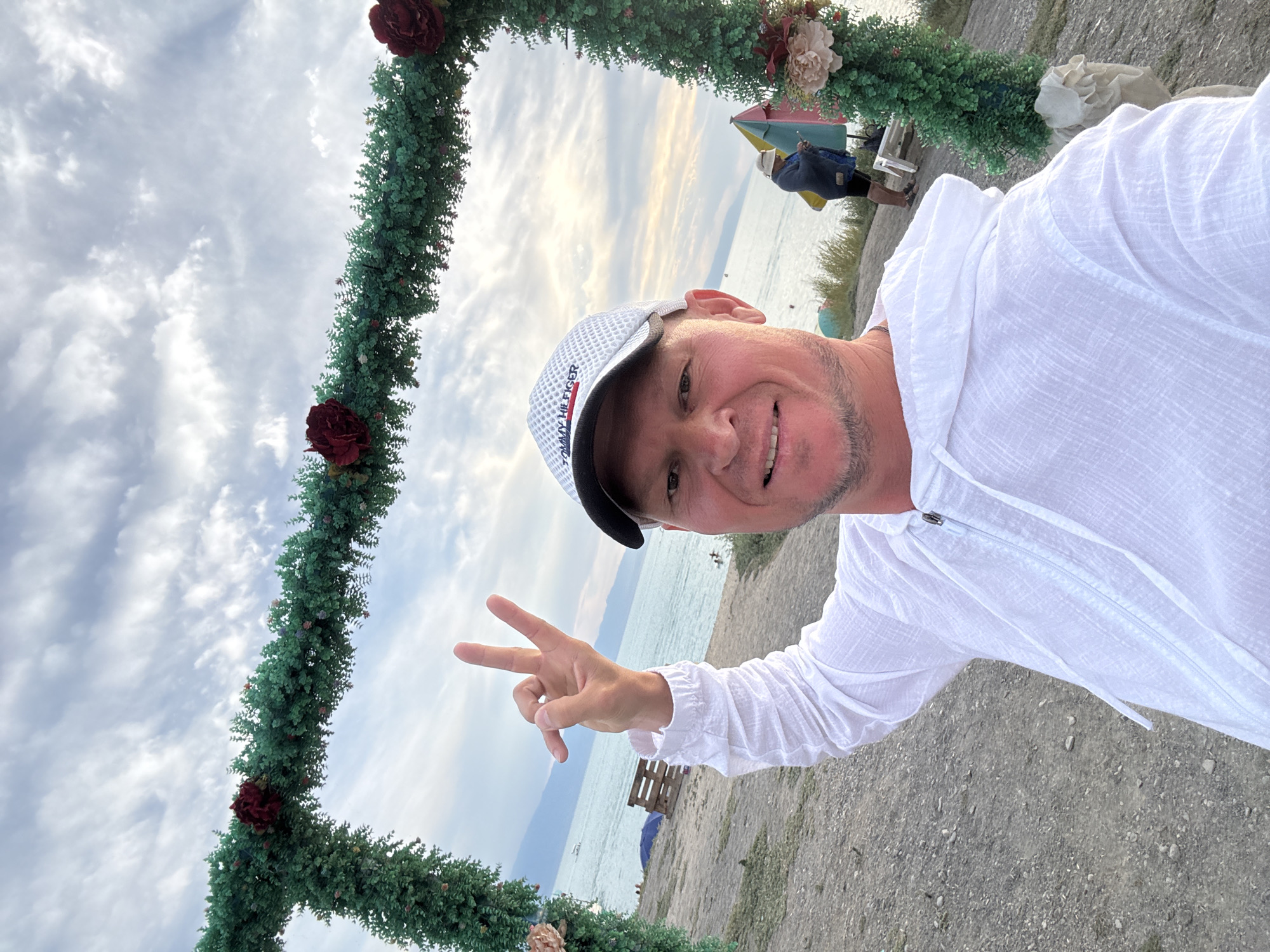 An outdoor, medium shot captures a smiling Caucasian man in his 40s posing for a selfie. He is wearing a white, hooded, linen shirt and a white baseball cap with "Tommy Hilfiger" written on it. His right arm is raised, and he is making a peace sign with his hand. The man is standing on a beach with a body of water and mountains in the background. To his left, a green archway decorated with red flowers is visible. In the background, other people are seen relaxing on the beach. The sky is cloudy with a mix of blue and grey. The location is likely Urdzhar, Kazakhstan, given the context of the query.