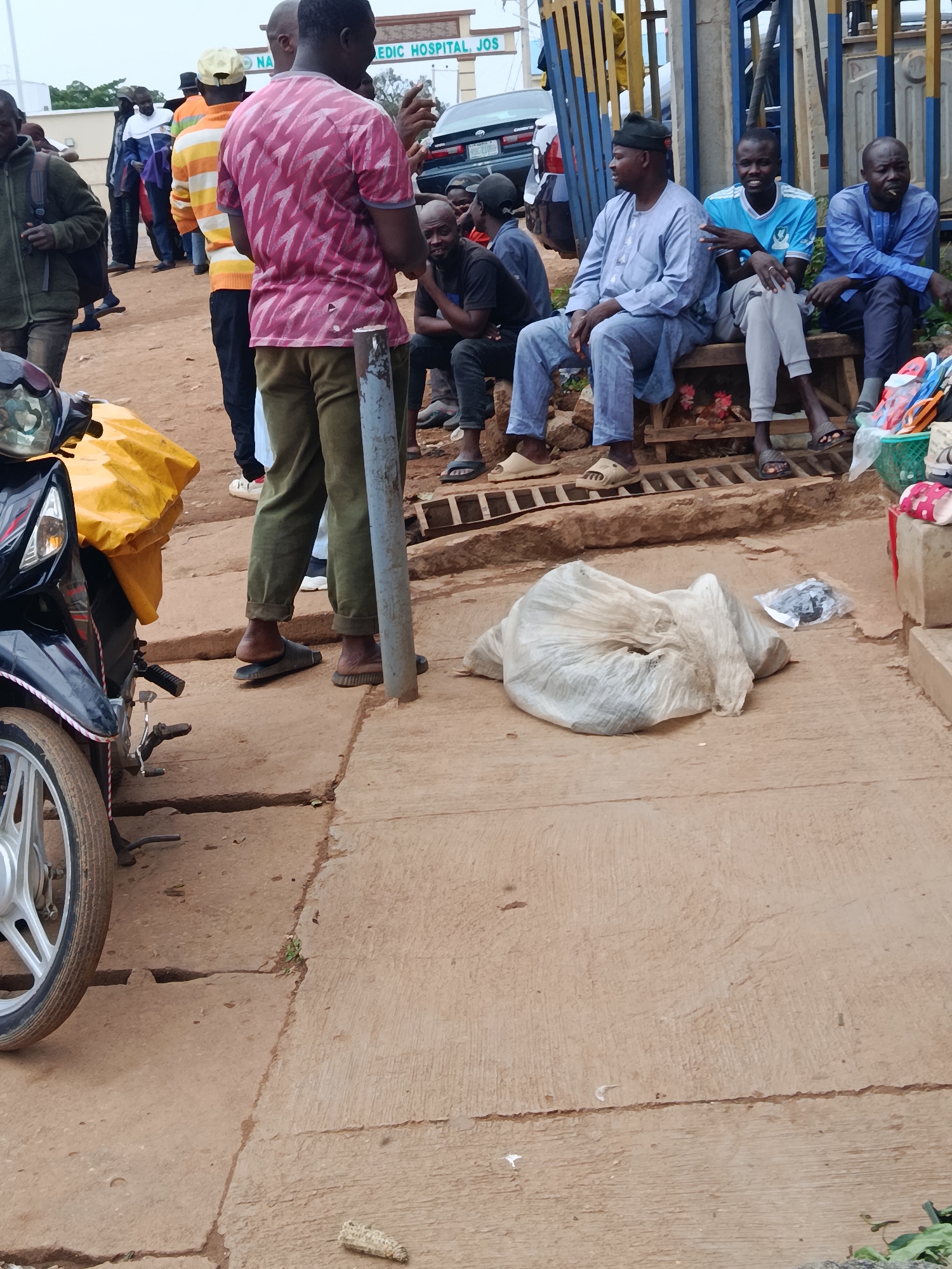 This image depicts a street scene in Jos, Nigeria, likely during daylight hours. A group of men are gathered, some seated on a wooden bench and others standing. One man in the foreground, wearing a pink and white patterned shirt and green pants, is gesturing with his right hand. To his left, a motorcycle with a yellow cover is parked.
The men seated on the bench are dressed in a mix of casual and traditional attire. One man wears a blue t-shirt and grey pants, while another is in a light blue traditional outfit. A third man on the bench is wearing a dark blue traditional garment. Some of the men are wearing sandals, while others have bare feet.
In the background, a sign reads "NA... EDIC HOSPITAL JOS," indicating the presence of a hospital. A dark-colored car is parked near the entrance. The ground is paved with concrete, and there are some cracks and unevenness. A large, white plastic bag filled with unseen contents is placed on the ground in the lower right portion of the image, alongside a smaller plastic bag with dark items. Further to the right, a basket containing colorful flip-flops is visible. The overall atmosphere appears to be one of casual gathering and waiting.