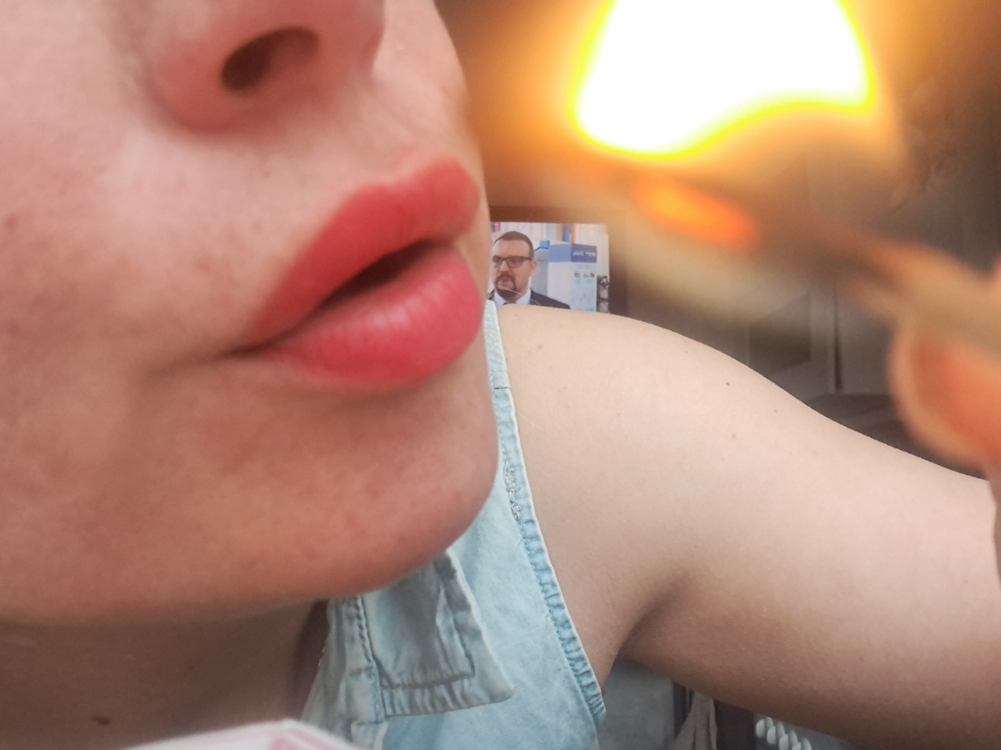 The image shows a close-up of a person's face, focusing on their nose and lips, which are painted with red lipstick. The person wears a light blue denim top. In the background, slightly out of focus, is a television screen displaying a man in a suit and glasses. There is a bright, blurry object on the right side of the image, possibly a light source. The image does not provide any information to place the location in the city of Elektrostal, Russia.