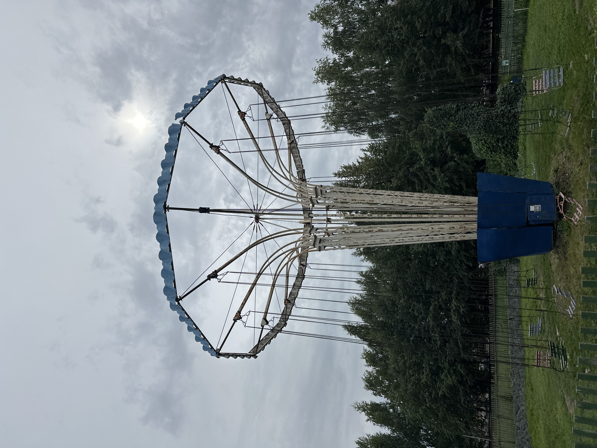 The image shows a vintage swing ride at an amusement park. The ride, which is partially rusted, consists of a central column from which chains extend to seats. The seats are empty, and the ride appears to be inactive. The background is filled with lush green trees and a cloudy sky with the sun peeking through. A wooden fence surrounds the area. The ride is located in the amusement park of Biysk, Russia.