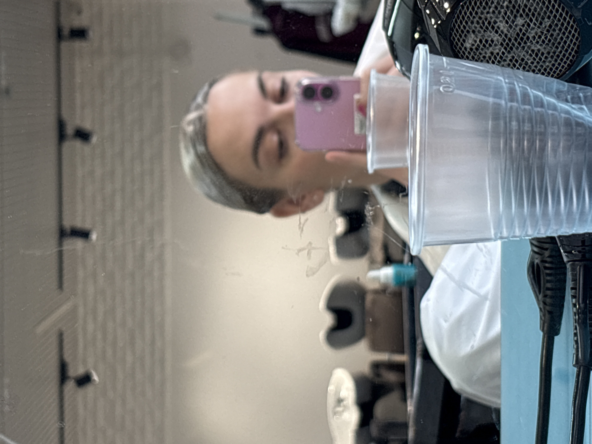 A person is holding a pink smartphone, taking a selfie in a mirror. The reflection in the mirror shows various salon equipment. The mirror has smudges and other marks on it. The person has light skin and light hair. A hair dryer and a stack of clear plastic cups are prominent in the foreground. The background shows seating and other items typically found in a hair salon. It is not possible to determine the location within Krasnoyarsk, Russia based solely on this image.