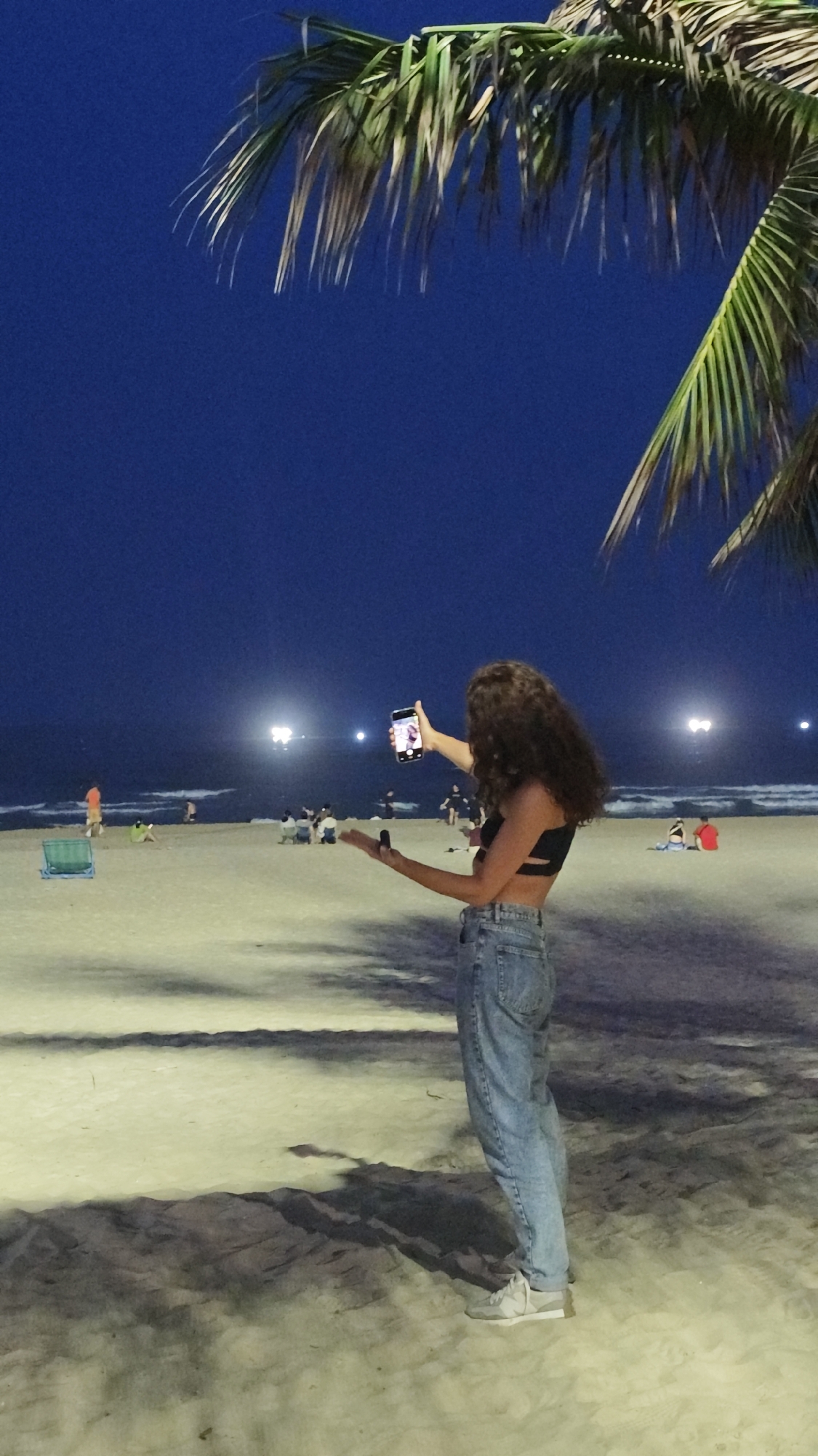 At night in Đà Nẵng, Vietnam, a woman stands on the beach taking a photo with her smartphone. She has curly hair, wears a black top and light-wash jeans, and faces the ocean. Her right arm is extended holding the phone, while her left arm is outstretched with an unidentifiable object in her hand. In the background, other people are seated or walking on the sand near the water. Lights illuminate the beach and the ocean horizon. A palm tree is partially visible in the upper right corner of the frame. Shadows of the tree and the woman are cast on the sand.