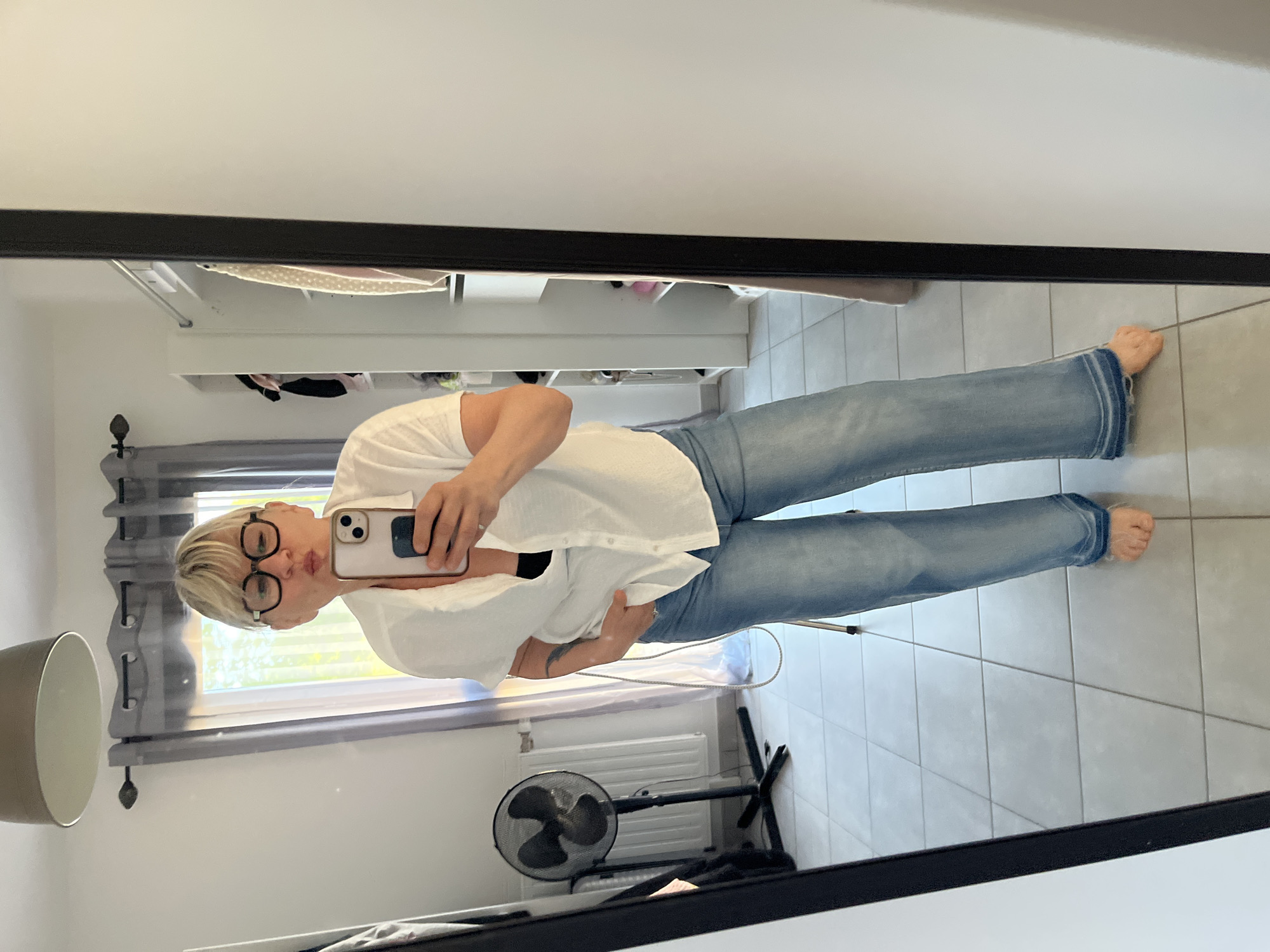 A woman is taking a selfie in a mirror. She is wearing glasses, a white shirt, and blue jeans. She is standing barefoot on a tiled floor. In the background is a window with curtains, a radiator, a fan, and some furniture. The location is Chambœuf, France.
