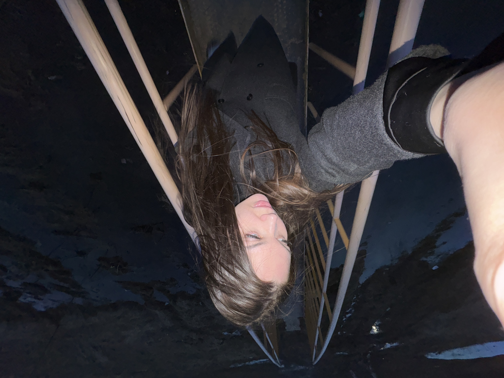 A photograph shows a woman in a gray coat, with long brown hair, posing in what appears to be a structure with light-colored metal beams or railings. The image has been rotated 180 degrees. The background shows what seems to be dark, possibly wet or icy ground. The location is identified as the city of Фоки, Russia.