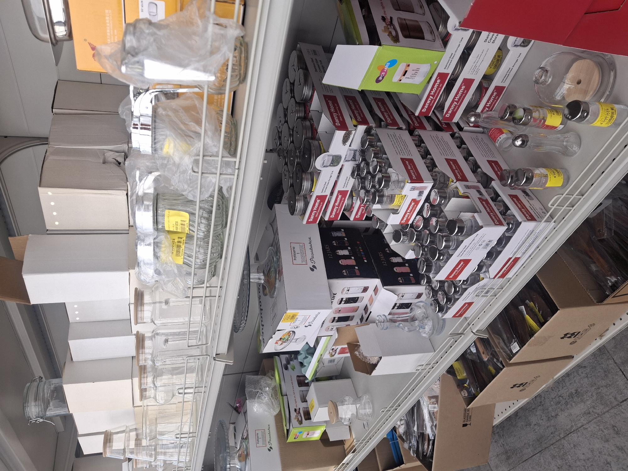 The image shows a retail store shelf stocked with various glass jars and containers. The shelves are metal, and the items are arranged in a structured manner. There are multiple types of glass jars visible, including some with wooden lids and others with metal lids, many of which are packaged in cardboard boxes. Some boxes are labeled with "Danny Home" and "Pasabahce," suggesting brand names. In the foreground, there are individual glass jars, some with yellow price tags. The background consists of more shelves filled with similar items and some larger cardboard boxes. The lighting appears to be artificial, typical of an indoor retail environment. Based on the visible text on some of the packaging, such as "Danny Home," the store is likely a kitchenware or home goods store. The presence of Cyrillic text on some of the packaging, combined with the brand names and the general appearance of the store, suggests this could be a store located in Tashkent, Uzbekistan, which has a significant Russian-speaking population and a market for goods from various international brands.