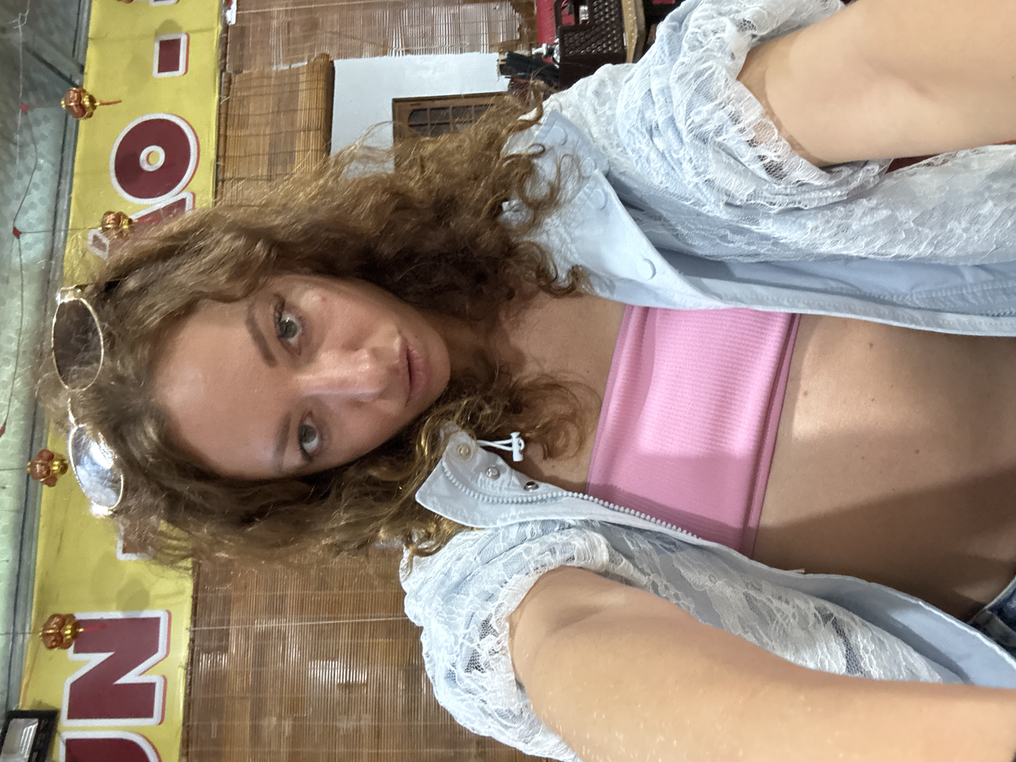 The image displays a close-up selfie of a female individual taken indoors in Trung Tam Ward, Vietnam. The individual has light skin, long, curly, reddish-brown hair, and is looking directly into the camera with a neutral expression. Dark sunglasses are perched on top of her head. She is wearing a light pink ribbed bandeau top under an open light blue collared jacket. The jacket features a zipper closure and white lace overlay details on the sleeves and parts of the body.
The background consists of a yellow banner or sign displaying large red characters, with "UN" visible on the left and a large "O" and "AO" visible on the right. Several small, red, decorative lantern-like objects are suspended from a light green ceiling. Below the banner, a section of light brown, horizontal bamboo blinds or screening is visible. To the right of the individual, a white wall and a wooden door with glass panels are present. Fragments of a dark, textured object and red fabric are discernible in the far right background.