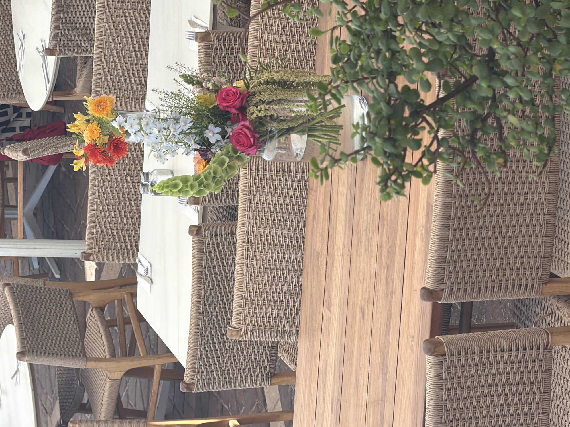 The image depicts an outdoor seating area at a restaurant, likely in Tel Aviv, Israel, given the presence of woven chairs and tables commonly found in Mediterranean settings. A wooden table occupies the foreground, with several woven chairs surrounding it. In the background, more tables and chairs are visible, suggesting a lively dining atmosphere. A floral arrangement in a glass vase sits on one of the tables, adding a touch of color and elegance to the scene. The overall impression is one of a relaxed and inviting dining experience.