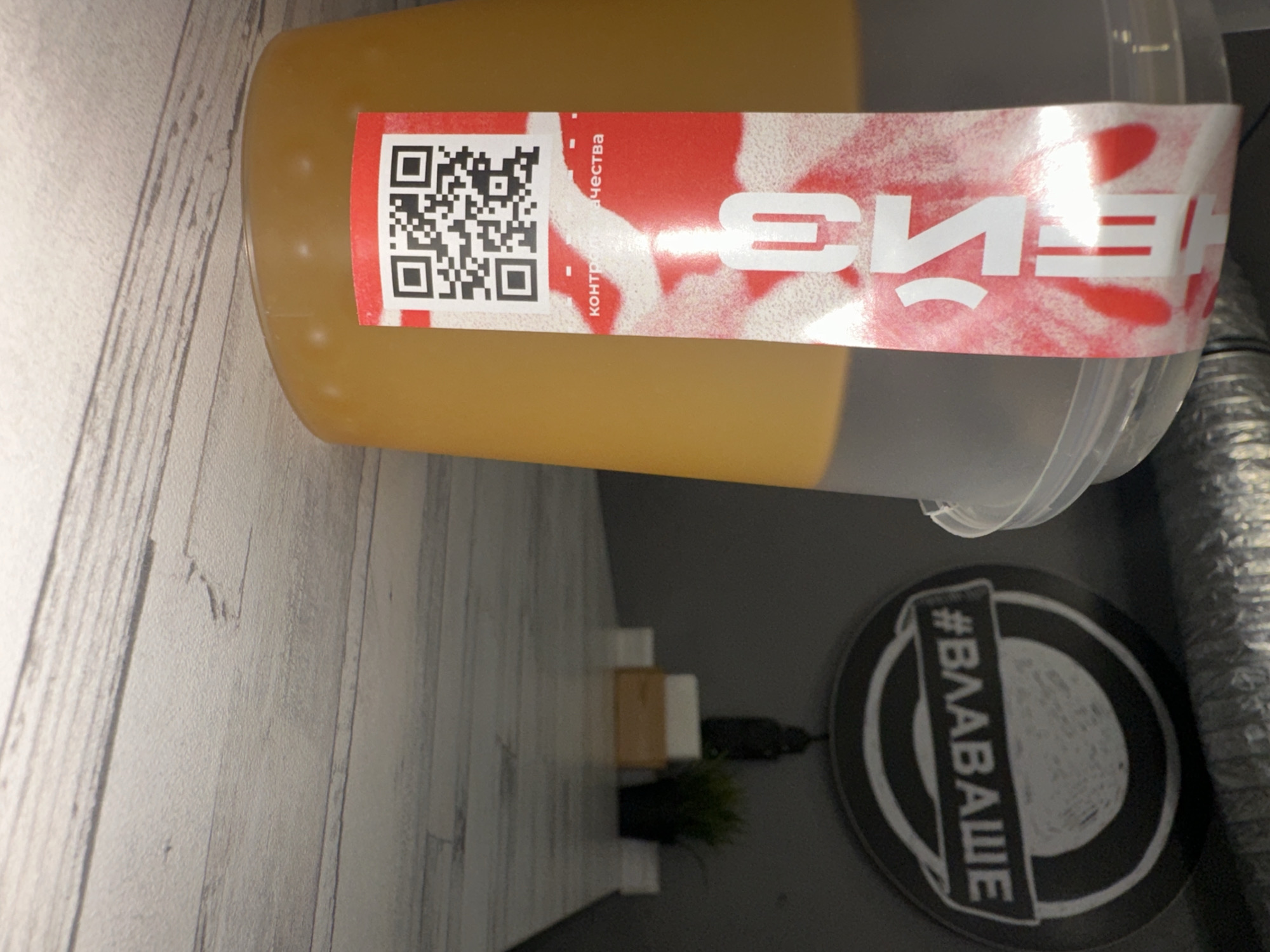 A clear plastic bottle or shaker, filled with a light amber or yellowish-brown liquid, is the central subject of this image, positioned at an angle. A red and white label is affixed to the bottle. On the left side of the label, a black QR code is clearly visible. To its right, the Cyrillic text "контроль качества" (quality control) is printed. Further to the right, large white Cyrillic characters are displayed against the red background, appearing mirrored or reversed, possibly indicating a brand name like "БЛЕНД" (BLEND).
The background features a light grey wall with a textured, wood-paneled appearance. Below the bottle, on a dark grey surface, a small decorative plant in a pot and a dark, indistinct object (possibly a figurine or statue) are visible. To the right, a black circular coaster with white outlines and white text is placed on the dark surface. The text on the coaster reads "#ВЛАВАШЕ" (#VLAVASHE).
The scene suggests an indoor setting, likely a home or office, with artificial lighting. No people or specific activities are depicted beyond the presence of the beverage container. The Cyrillic text on the label and coaster is consistent with the provided location context of Chaikovsky, Russia.