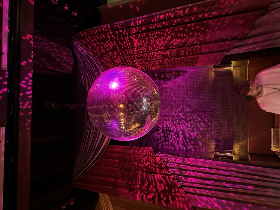 A disco ball hangs suspended in a dimly lit space, casting magenta and pink light across various surfaces. The light reflects on a textured floor and a tiered, patterned ceiling. Curtains drape around the ball, adding to the atmosphere. A figure stands to the right, possibly observing the scene. Overall, the image conveys a sense of nightlife and a celebration.
