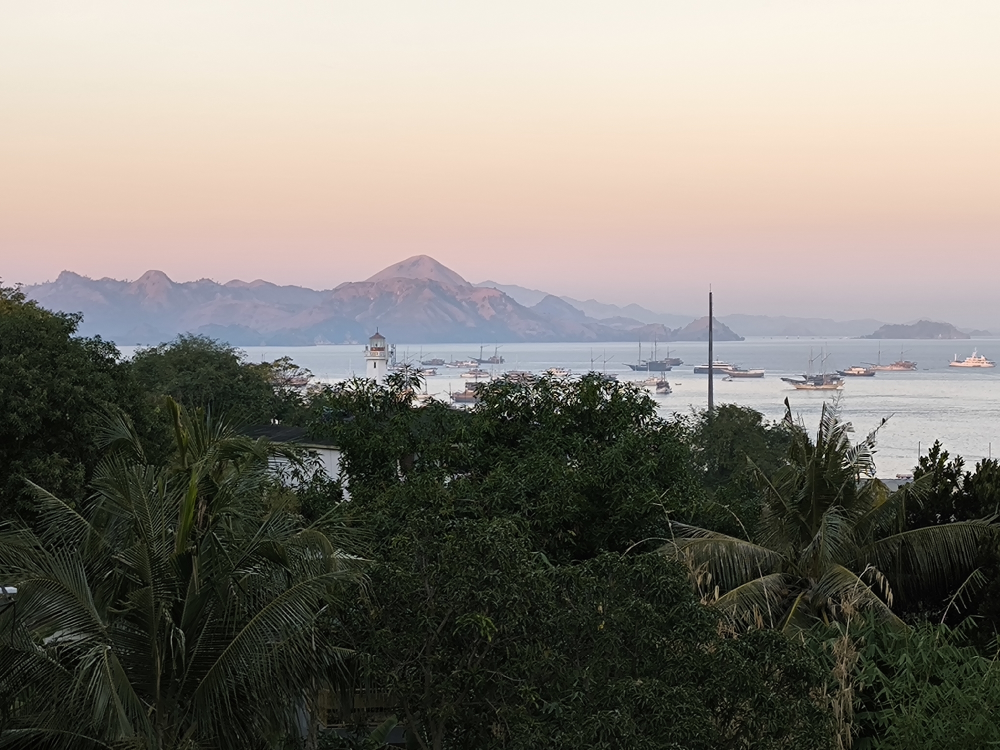 The image depicts a panoramic view of a harbor in Labuan Bajo, Indonesia, at dusk. In the foreground, dense green foliage, including palm trees, partially obscures the view of a body of water filled with numerous boats and ships of various sizes, some appearing to be traditional wooden vessels and others more modern yachts. A white lighthouse stands prominently on a hill overlooking the harbor. In the background, a range of layered mountains under a soft, gradient sky that transitions from a pale orange to a light purple. The overall atmosphere is serene and tranquil, indicative of the end of the day.