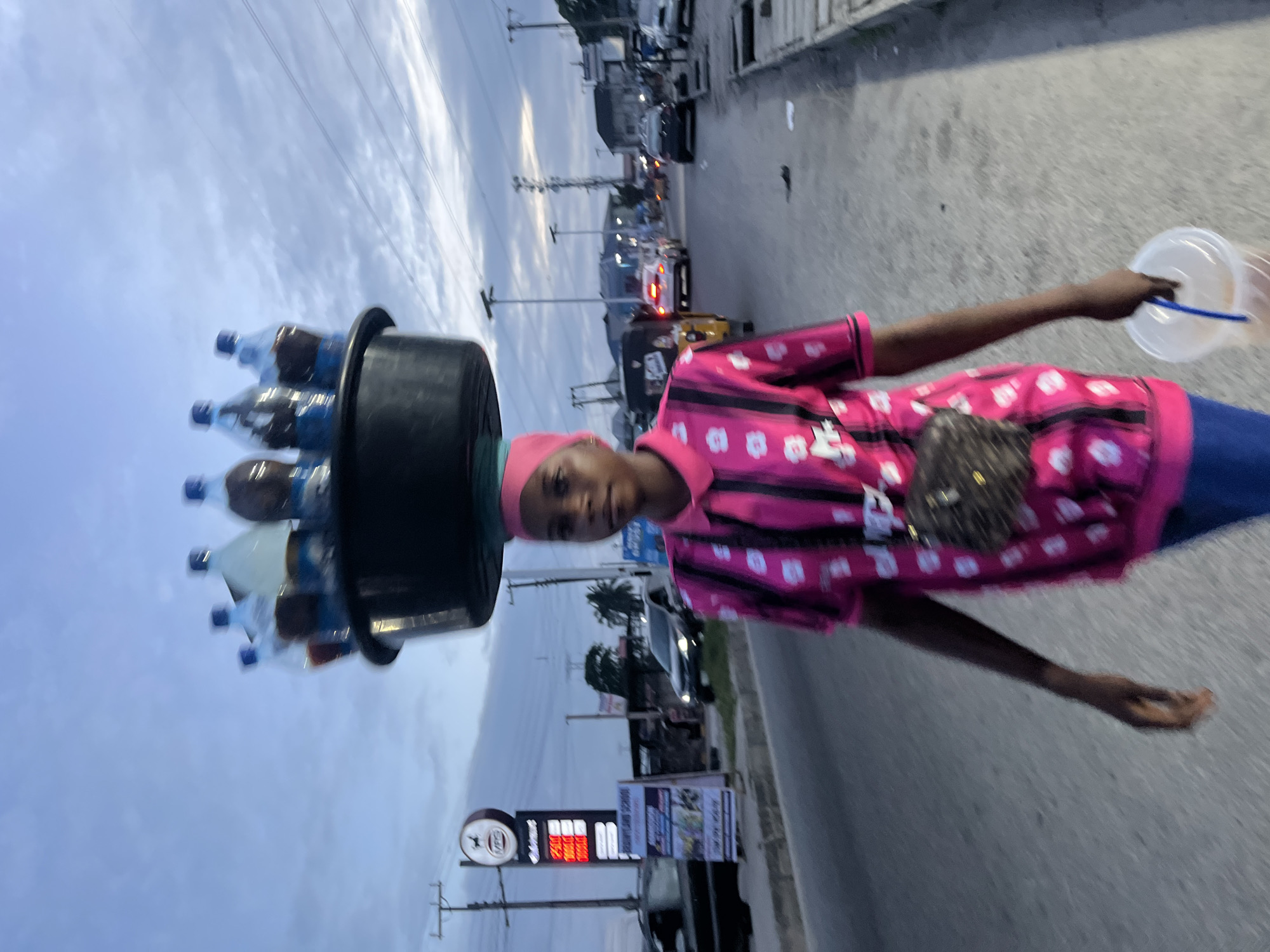 A young woman walks on the side of a busy road in Ado, Nigeria, carrying a large black basin filled with several plastic bottles of drinks on her head. She wears a bright pink, patterned shirt and blue pants, with a small dark purse slung over her shoulder. In her right hand, she holds a clear plastic bucket with a blue handle, containing a liquid that appears to be a beverage. The road is paved and bordered by a concrete walkway and drainage system. Several vehicles, including cars and a tricycle, are visible on the road, indicating a bustling urban environment. In the background, there are buildings, streetlights, and power lines, suggesting a typical commercial or residential area. The sky is overcast with a mix of blue and gray clouds, indicating either dawn or dusk. A gas station sign with illuminated price displays is prominent on the left side of the road. The overall scene captures a moment of daily commerce and transportation in a Nigerian city.