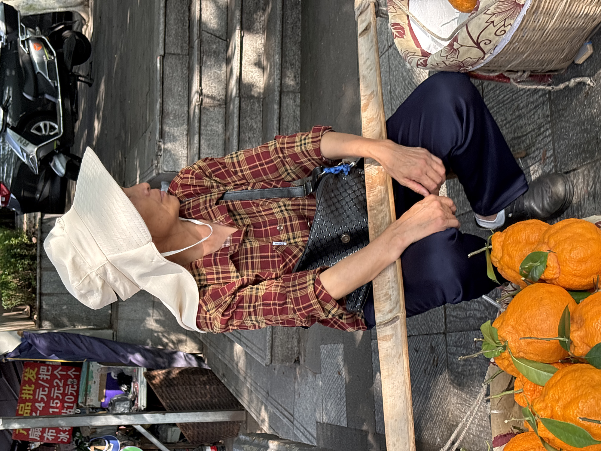 A person wearing a wide-brimmed hat and a plaid shirt sits on a wooden plank in what appears to be Yuzhong District, China. They are looking to the right. In front of them, an assortment of bumpy, orange-skinned fruits, possibly Japanese citrus, are arranged in a woven basket and on the ground. The person is wearing dark pants and black shoes. Behind them, a stone staircase leads upwards, and a scooter is parked on a paved surface. A sign with Chinese characters and pricing information is visible to the left. The scene is illuminated by sunlight, casting shadows on the ground and steps.