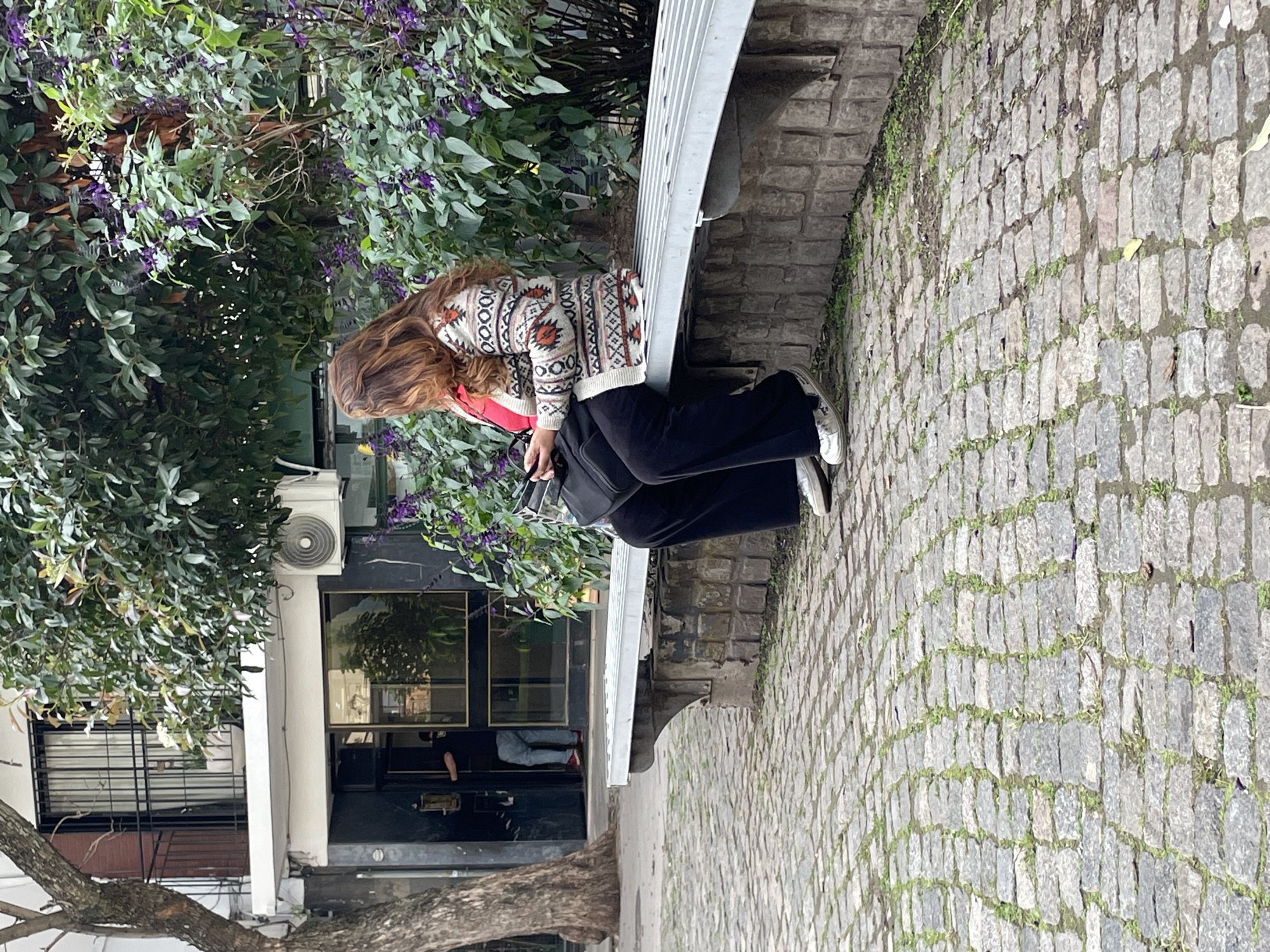 A medium shot captures a woman sitting on a stone bench in what appears to be an urban setting in Buenos Aires, Argentina. The woman has long, wavy brown hair and is wearing a cream-colored sweater with intricate red, black, and white geometric patterns. She is dressed in black wide-leg pants and white sneakers, with a red bag strap visible on her left shoulder. A dark bag rests on her lap as she looks downwards.
Behind the woman is a dense bush with green leaves and clusters of small purple flowers. To her right, a cobblestone pathway, typical of many streets in Buenos Aires, stretches into the background, with patches of green moss growing between the grey stones. The bench itself features a metal slatted backrest.
On the left side of the frame, partially obscured by a tree trunk in the foreground, is the facade of a building or shop. It features large dark windows reflecting the surroundings, and an outdoor air conditioning unit is mounted high on its wall. A person is faintly visible inside the building through one of the windows.
The scene conveys a calm, daytime atmosphere, possibly under an overcast sky or during late afternoon, as there are no harsh shadows. No discernible text is visible in the image.