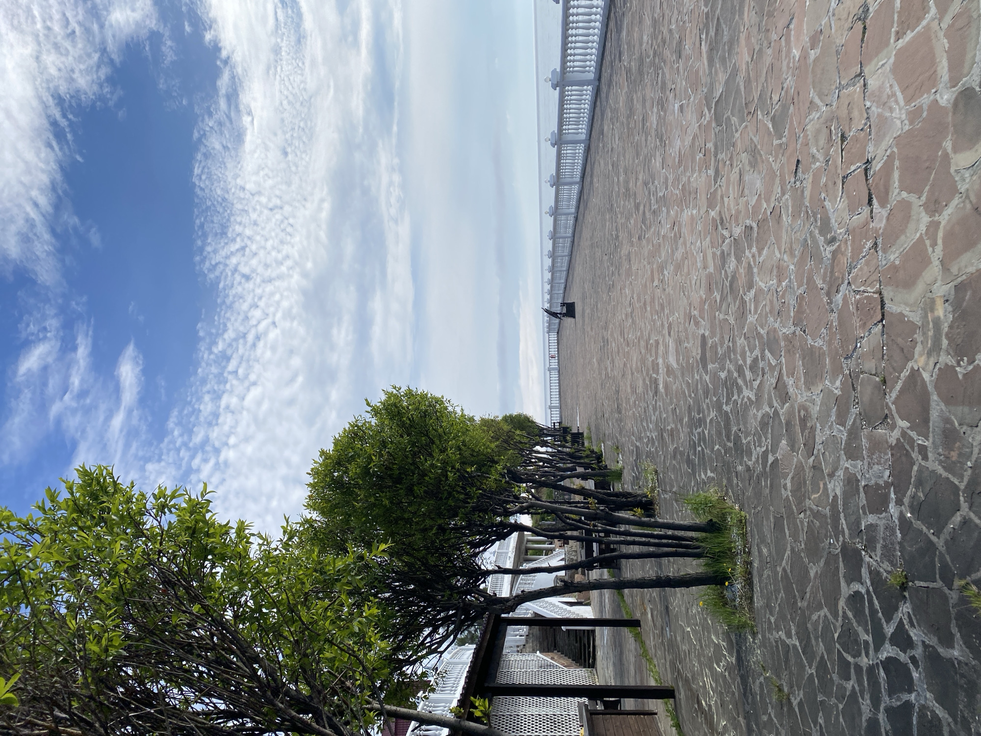 The image depicts a paved walkway lined with young trees and a white railing along the edge of a body of water, likely Lake Baikal, in Listvyanka, Russia. The walkway is constructed of irregular, flat stones. On the left side of the walkway, a white building with ornate railings and a covered area is visible. A series of young trees with bright green leaves are planted in a row along the edge of the walkway, separating it from the building. In the distance, along the railing, a lone dark-colored bench or statue is visible. The sky is partly cloudy with a mix of blue sky and white, wispy clouds. The water stretches out to the horizon, appearing calm. The scene is tranquil and appears to be an outdoor recreational area. There are no people visible in the image.