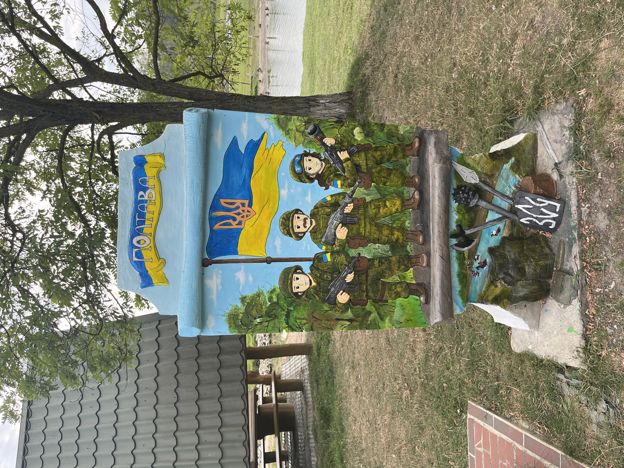 The image shows a sculpture depicting three soldiers in military uniform. The sculpture is situated outdoors in a grassy area with trees and a body of water in the background. The soldiers are painted in a cartoonish style, holding rifles and wearing helmets. The sculpture is adorned with the Ukrainian flag and the word "ПОЛТАВА" (Poltava) written in yellow and blue. The lower part of the sculpture features a painting of a river with some figures and a bucket, as well as what appear to be medieval weapons. A number "309" is painted in white on a dark background at the base. To the left of the sculpture is a picnic area with a covered table and benches, and a brick pathway. The overall scene suggests a public space, likely a park or recreational area, with patriotic artwork.