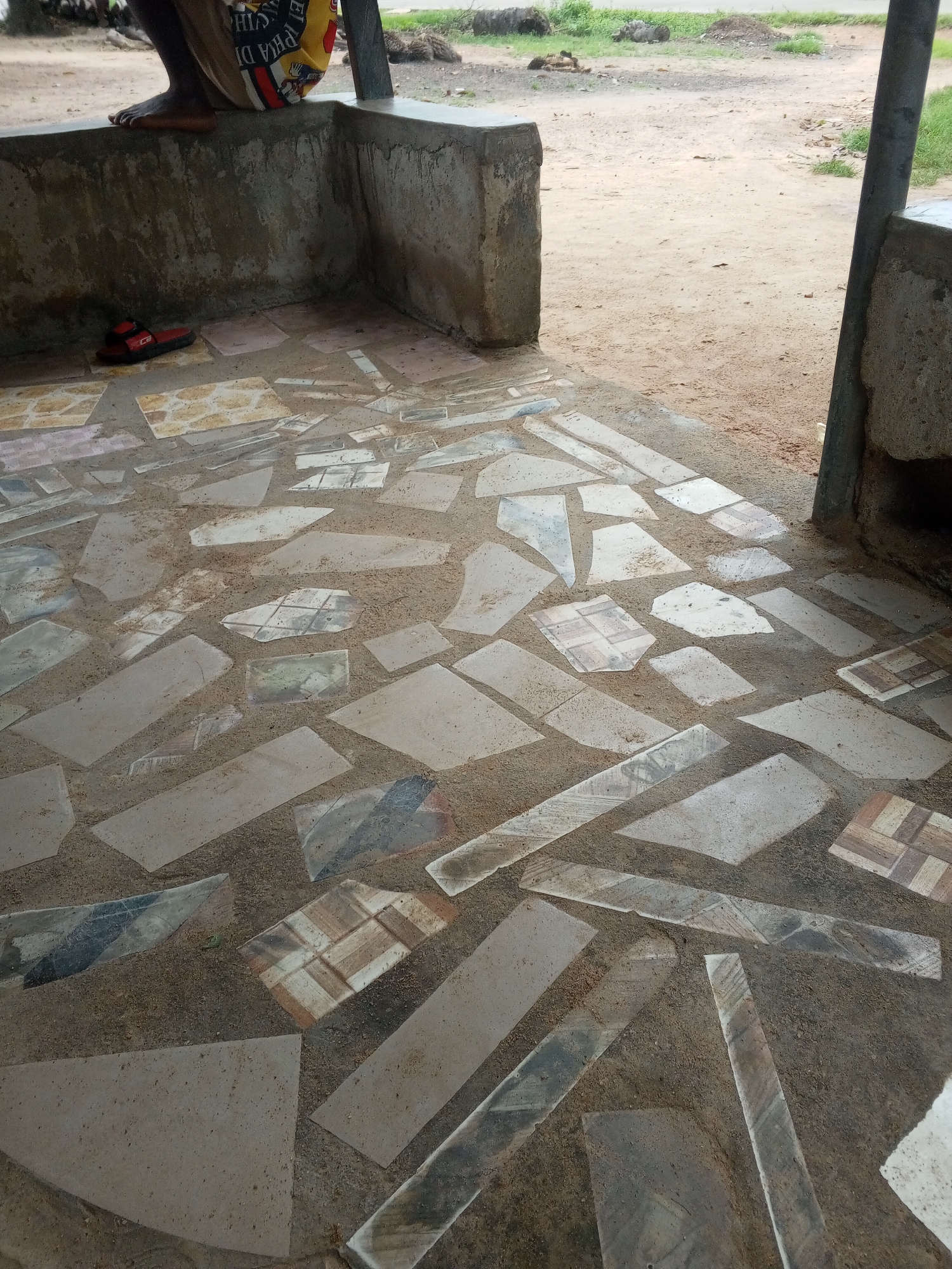 The image shows a close-up view of a mosaic tiled floor, likely a patio or walkway. The tiles are irregular in shape and size, with a variety of colors and patterns, including plain beige, grey, and some with intricate designs resembling wood or marble. They are embedded in a rough, sandy grout. To the left of the tiled area, a person's lower legs and feet are visible, with one foot resting on a low concrete wall. The person is wearing a colorful patterned cloth around their waist. On the ground near the wall are a pair of red sandals. The setting appears to be outdoors, with a sandy, dirt ground visible in the background. The lighting suggests it is daytime, possibly morning or afternoon, with shadows indicating sunlight. The overall impression is of a rustic, possibly unfinished outdoor space in a rural or semi-rural location in Gindin Dutse, Nigeria. There is no visible text in the image.