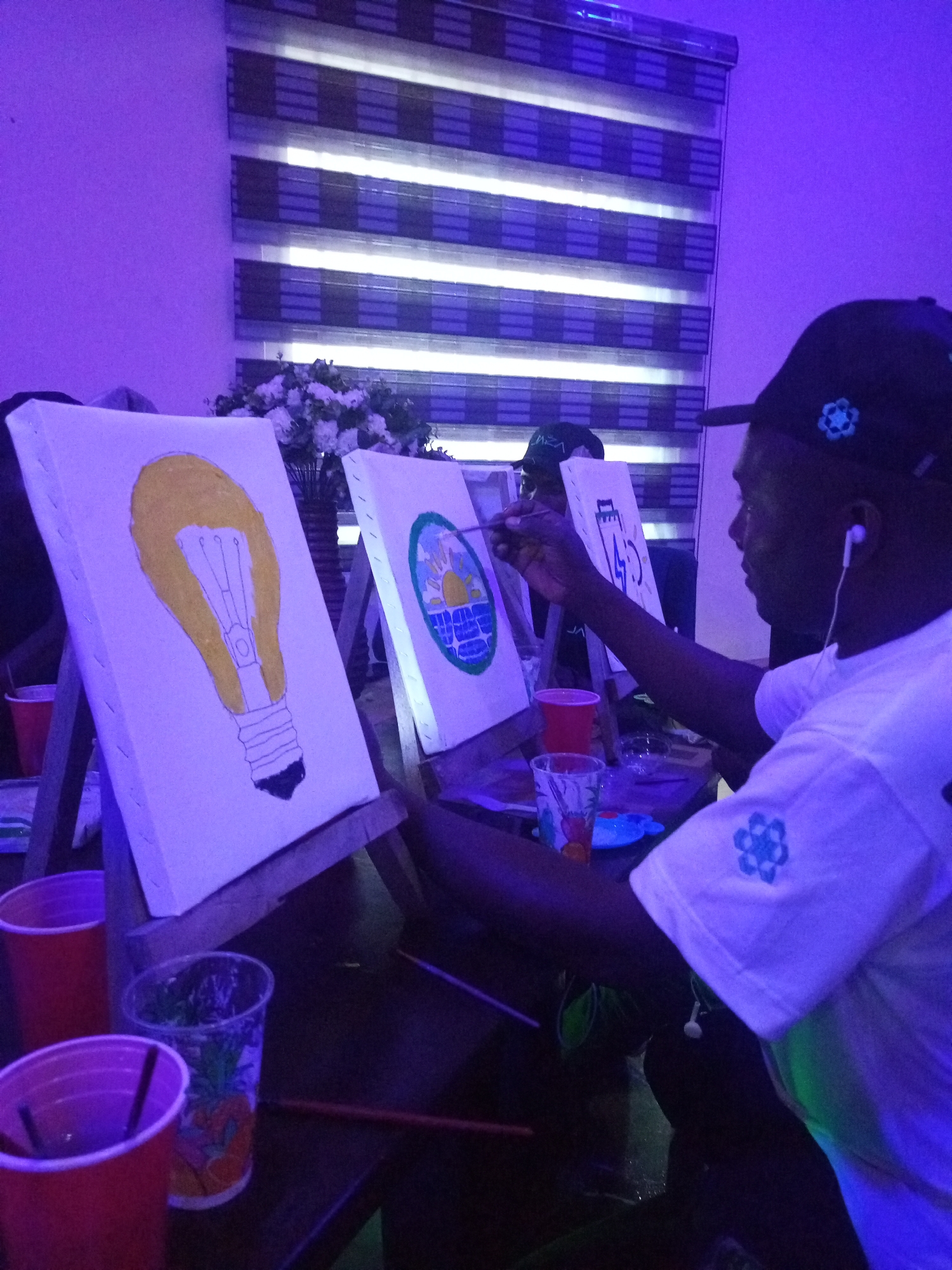 The image captures a group of people engaged in a painting session, likely an art class or social gathering, in Ado Ekiti, Nigeria. The lighting is predominantly purple, casting an atmospheric glow on the scene, suggesting it might be evening or a dimly lit indoor space.
The primary subjects are individuals focused on their canvases. One person in the foreground, wearing a white t-shirt and a black baseball cap adorned with a blue floral emblem, is actively painting. They have earbuds in, indicating a personal soundtrack or focus. Their concentration is evident as they hold a paintbrush, actively working on a canvas positioned on an easel.
To the left, another canvas displays a yellow lightbulb with a black base, also on an easel. Further back, a third canvas shows a depiction of a sunset over water, enclosed in a circular frame. In the background, a vase of white flowers adds a decorative touch. The setting appears to be an indoor space with horizontal blinds on a window or wall behind the participants.
Several plastic cups, predominantly red and clear with colorful patterns, are visible on the table, likely containing drinks. The presence of easels, canvases, and paintbrushes clearly indicates that painting is the central activity. The overall mood seems relaxed and creative, with individuals immersed in their artistic endeavors. The purple lighting creates a modern, perhaps even playful, ambiance for the event. There is no visible text in the image.