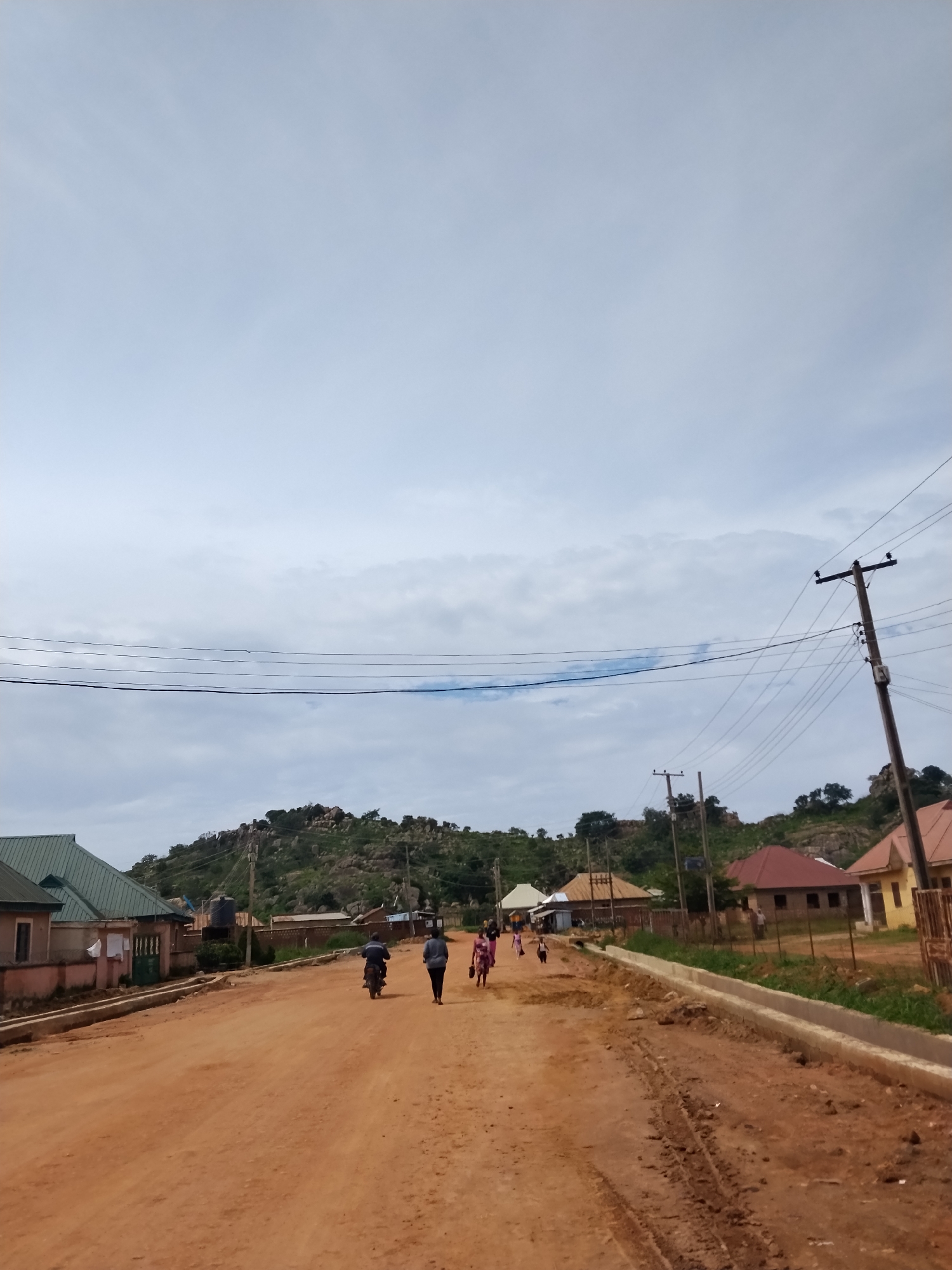 The image depicts a street scene in Laminga, Nigeria. The foreground is a dirt road with some tire tracks and a newly constructed concrete drainage channel along the right side. In the middle ground, several people are walking or riding a motorcycle. On the left side of the road, there are houses with green and pink facades and metallic roofs. On the right side, there are houses with red and yellow roofs. In the background, a hilly, rocky landscape is covered with vegetation. Several utility poles with wires are visible throughout the scene, indicating the presence of electricity infrastructure. The sky is overcast with clouds.