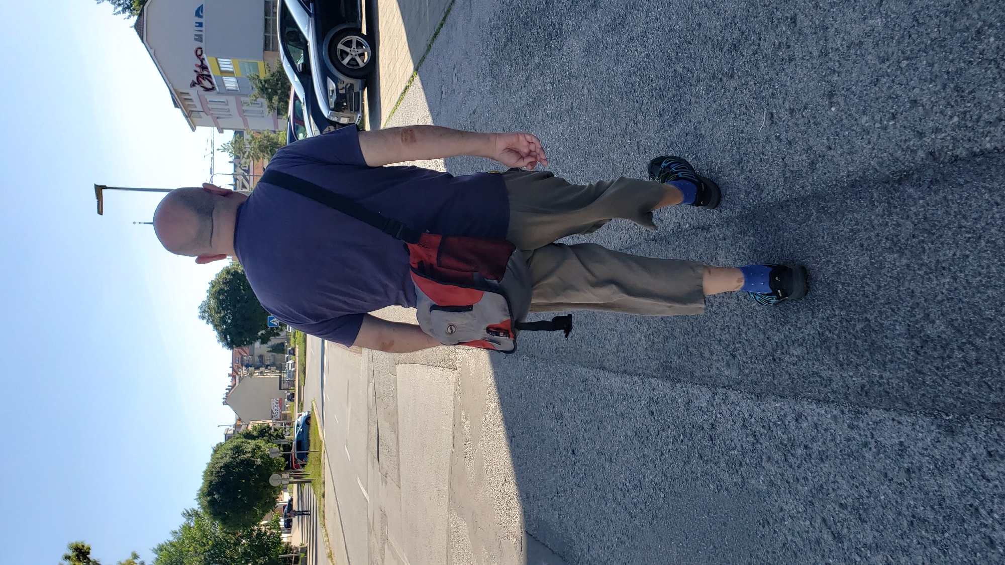 A bald man wearing a navy t-shirt, khaki pants, blue socks, and black sandals walks away from the camera on an asphalt road. He carries a red and grey shoulder bag across his chest. The street is lined with buildings and trees, and cars are parked on the right side of the road. The scene is set in the city of Prešov, Slovakia.