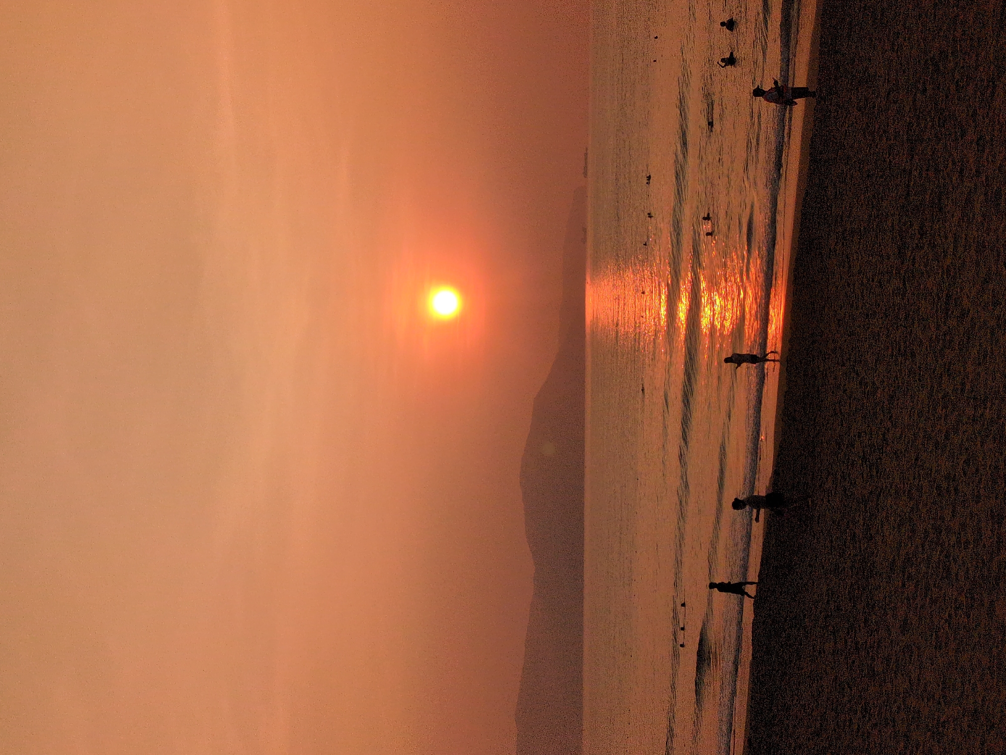 The image shows a sunset scene on a beach in South Quy Nhon Ward, Vietnam. The sun is setting over the ocean, casting a warm orange glow across the sky and water. Several people are visible on the beach and in the water, appearing as silhouettes. The beach consists of sand. The foreground is a darker area, presumably the sand closer to the viewer. The water reflects the light of the setting sun. A hill or mountain is visible in the distance.