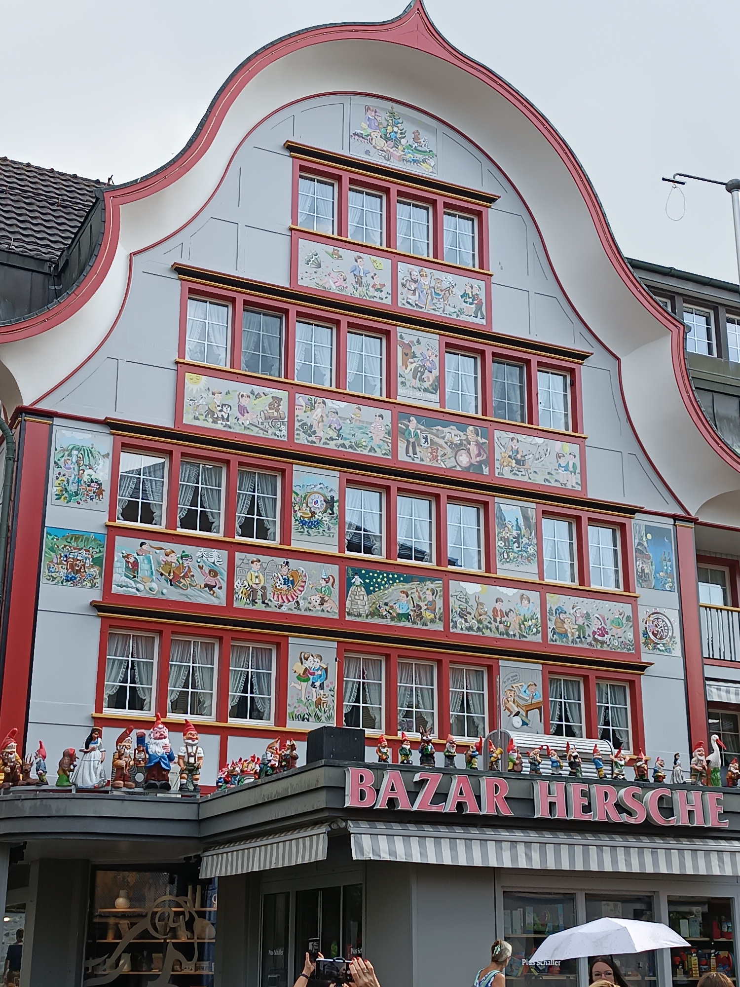 In Appenzell, Switzerland, a detailed facade of a building is captured, characterized by a grey exterior with ornate red trim and numerous decorative panels. These panels depict various scenes, many of which feature stylized figures and children engaged in activities, often with a whimsical or folkloric theme. The windows are numerous and are framed by the same red trim, with white curtains visible behind the glass.
At the lower section of the building, a prominent sign reads "BAZAR HERSCHE" in large pink lettering. Below the sign, a striped awning extends outwards, and a shop entrance is visible, showcasing merchandise.
Along the roofline of the shop, above the "BAZAR HERSCHE" sign, a collection of garden gnomes and other figurines are arranged. These figures vary in size and detail, with many wearing traditional gnome attire such as red pointed hats. Among the gnomes, there are also figures of a woman in a white dress and a stork.
The overall impression is of a brightly decorated building with a commercial establishment at its base, set against an overcast sky. The scene is a street-level view, suggesting a public area with pedestrian activity, though the focus is primarily on the architecture and the display of the shop.
