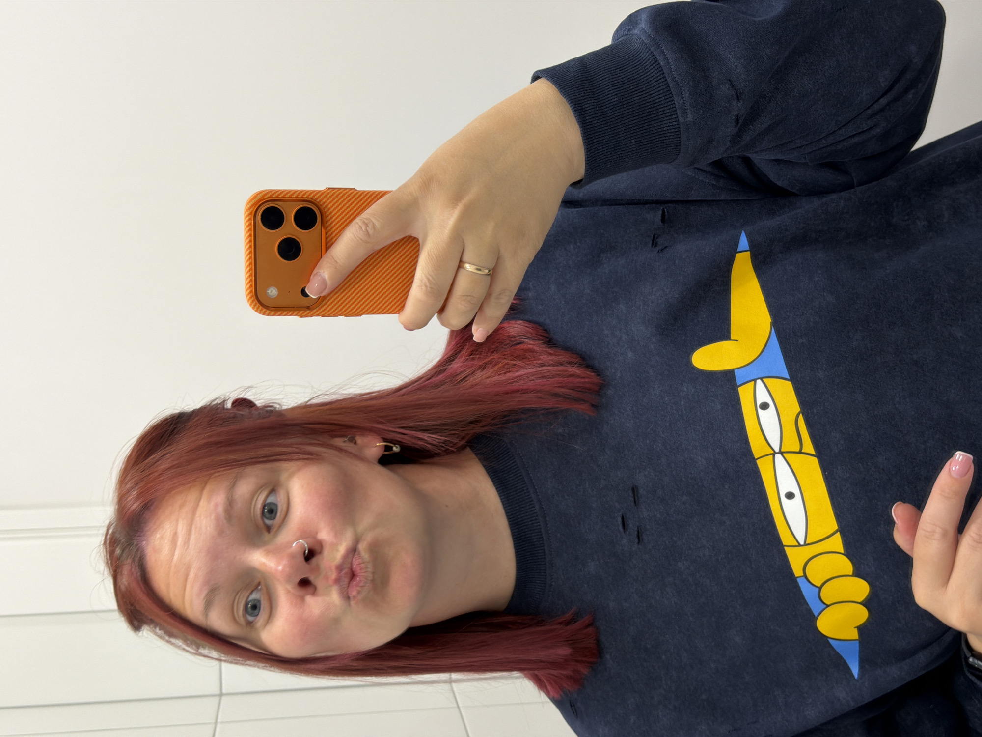 A person is taking a selfie in what appears to be a bathroom setting. The person has reddish-brown hair, blue eyes, and a nose piercing. They are puckering their lips and looking directly at the camera, which is an orange-cased cell phone held in their right hand. The person is wearing a dark-colored sweatshirt with a cartoon graphic on the front. There are some small rips or distress marks on the sweatshirt. A plain gold ring is visible on the ring finger of their right hand. The background consists of plain white walls, possibly tiled. The location appears to be a bathroom, but no specific information or details indicate that it is in the city of Chaikovsky, Russia.
