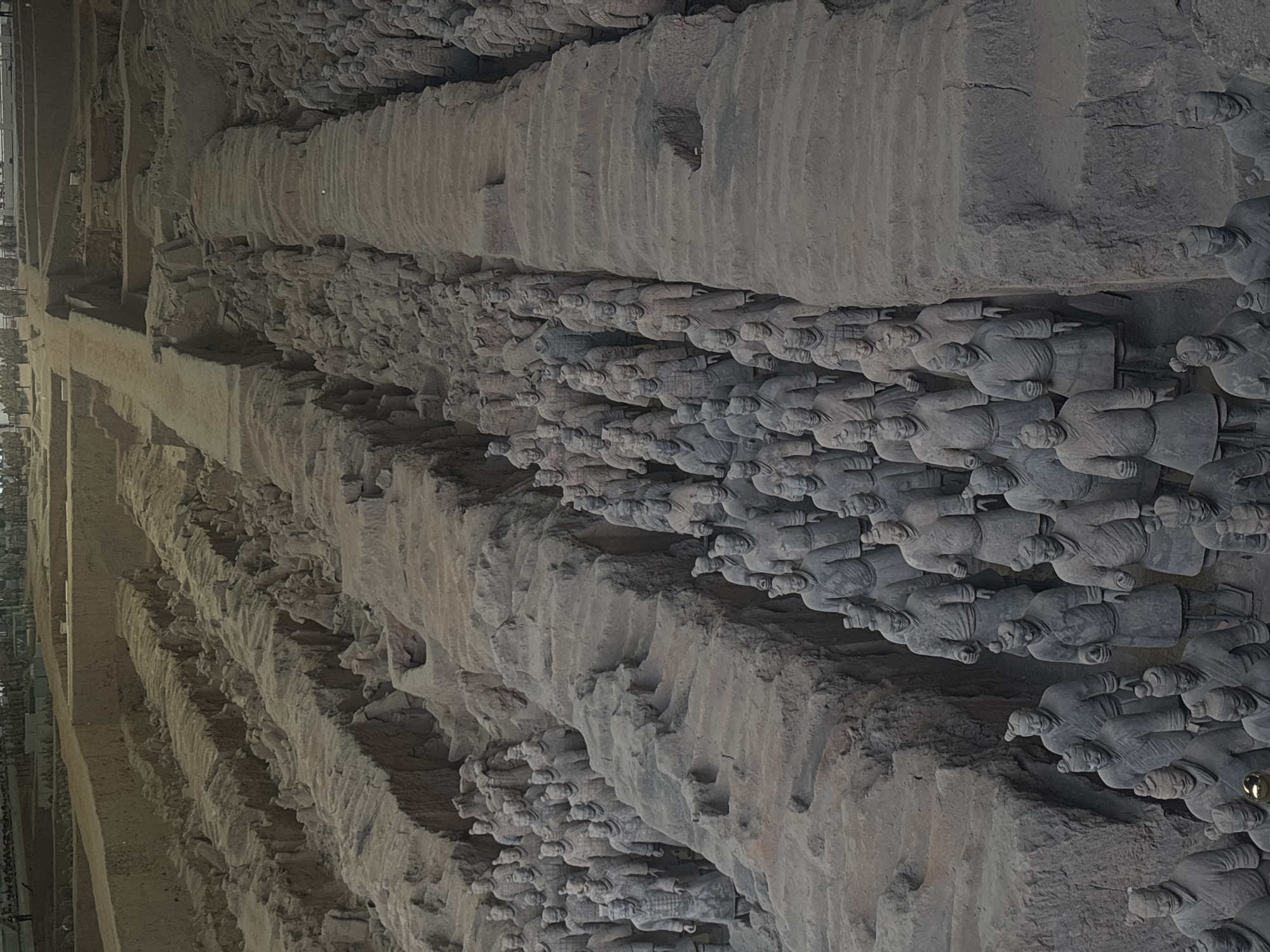 The image shows a detailed view of the Terracotta Army in Lintong District, China. The scene depicts a vast excavation site with rows upon rows of life-sized terracotta warrior statues arranged in battle formations within trenches. The figures are made of clay and vary in height, attire, and hairstyle, suggesting different ranks within the army. Many statues are in various states of preservation, with some appearing more intact than others, and the earth and debris around them indicate ongoing archaeological work. The overall impression is one of immense scale and historical significance, with the statues appearing to march in an organized and formidable manner. The lighting suggests an indoor or covered environment, likely a museum or protected excavation pit.