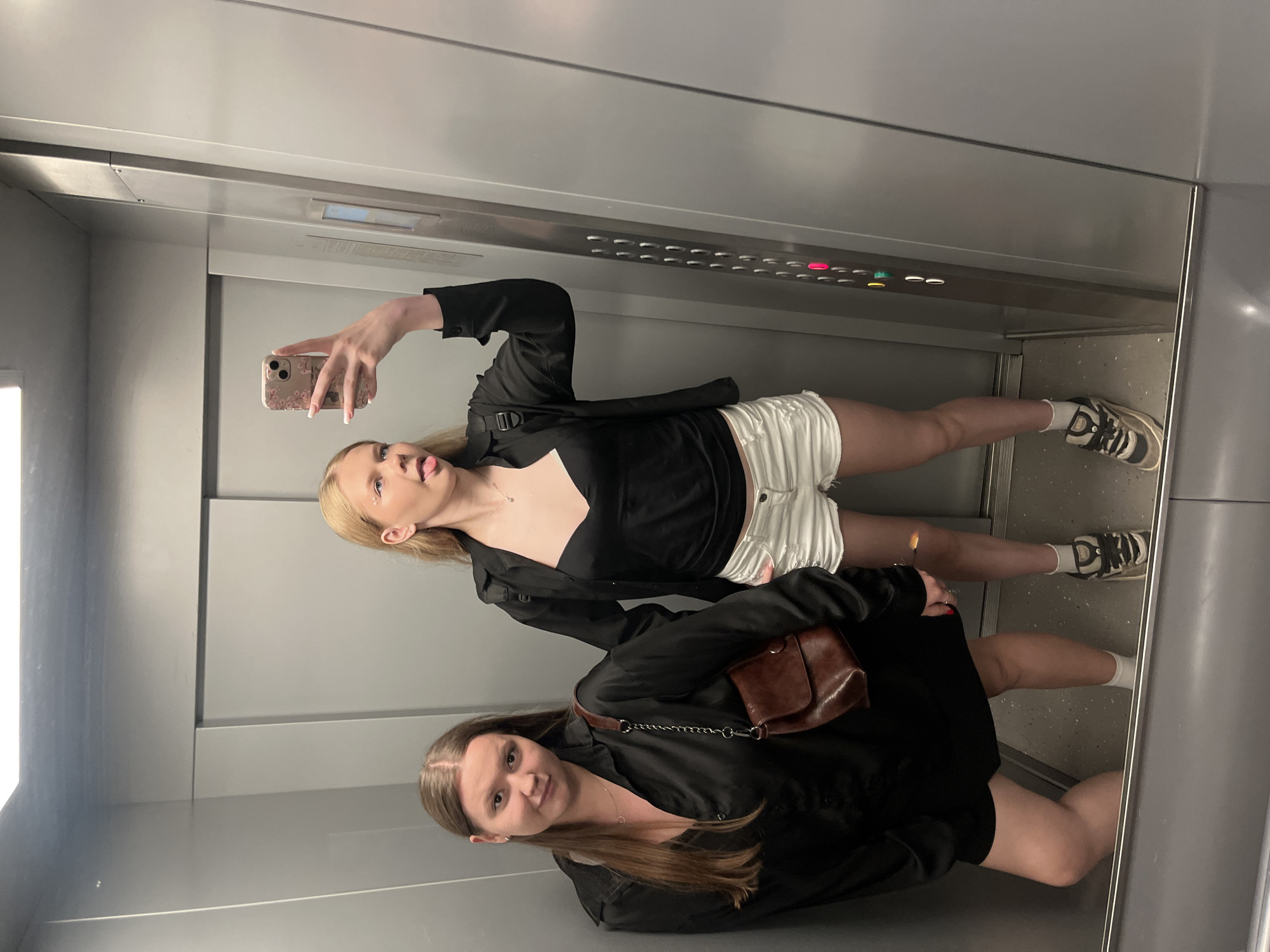 Two young women are taking a selfie in an elevator. The woman on the left is wearing a black oversized button-down shirt, black shorts, and white socks. She has long brown hair and is carrying a brown leather crossbody bag. The woman on the right is wearing a black camisole, white denim shorts, and white socks with sneakers. She has blonde hair and is sticking her tongue out at the camera. The elevator has gray walls and a metal control panel with illuminated buttons. The floor of the elevator is speckled gray. The scene appears to be in a modern building. There is no specific indication of the location being in the city of Perm, Russia.