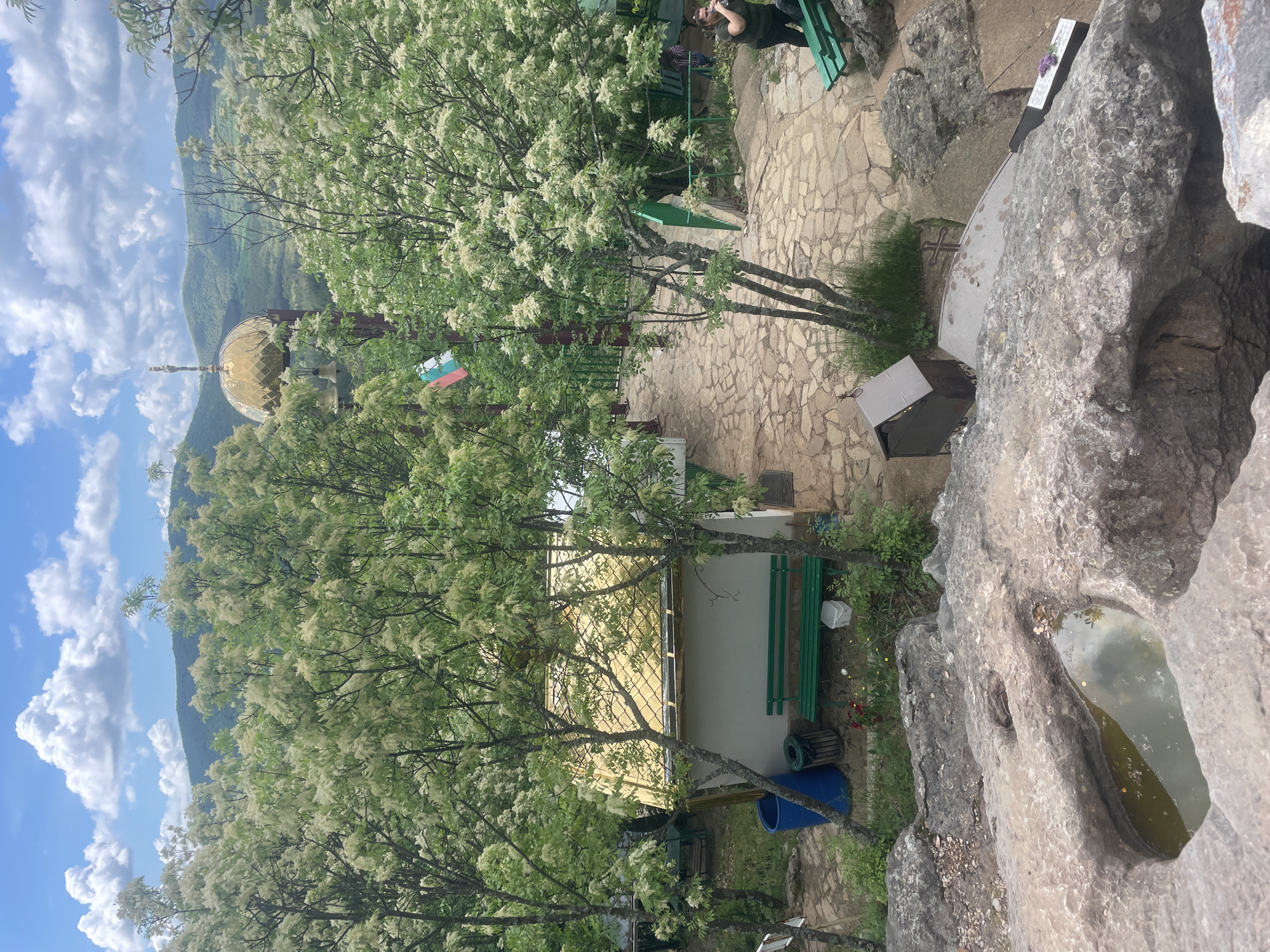 The image shows a view from a rocky outcrop in Novo Selo, Bulgaria. A golden-domed structure is visible among trees bearing white blossoms. The Bulgarian flag is also seen. Below, a building with a light-colored facade stands adjacent to green benches. Paved stone is visible along with various containers. In the foreground, a natural rock pool contains water, reflecting the sky.